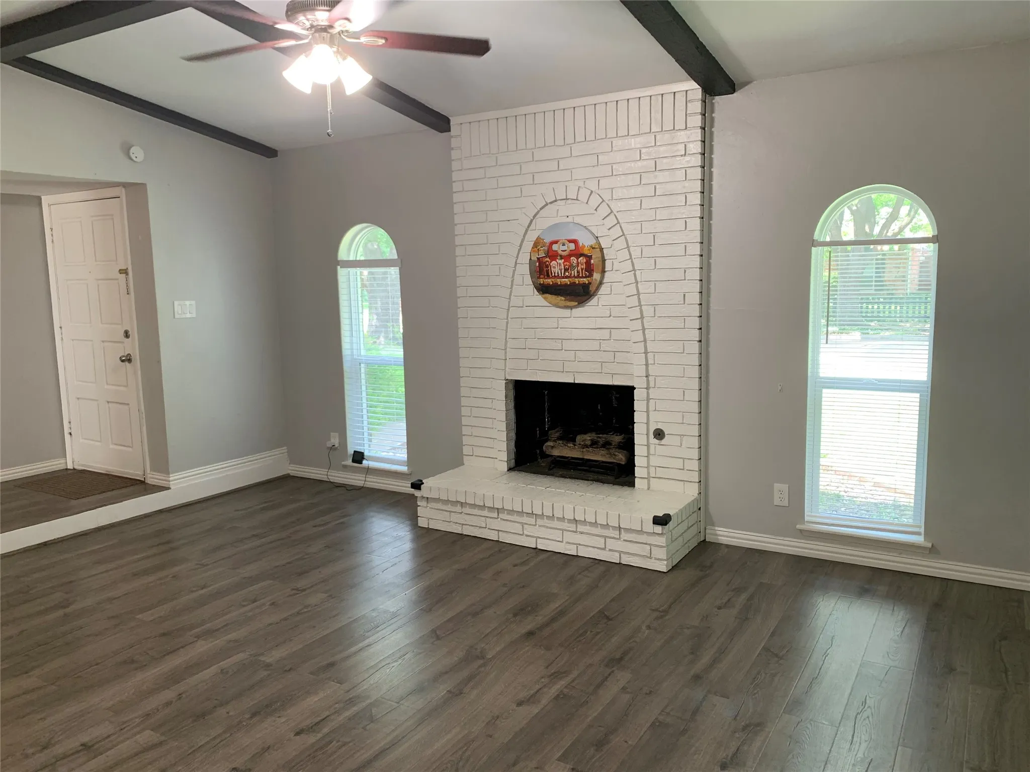 Living area featuring a painted brick fireplace, wood-finish flooring, two arched windows, a ceiling fan with integrated lighting, and exposed ceiling beams