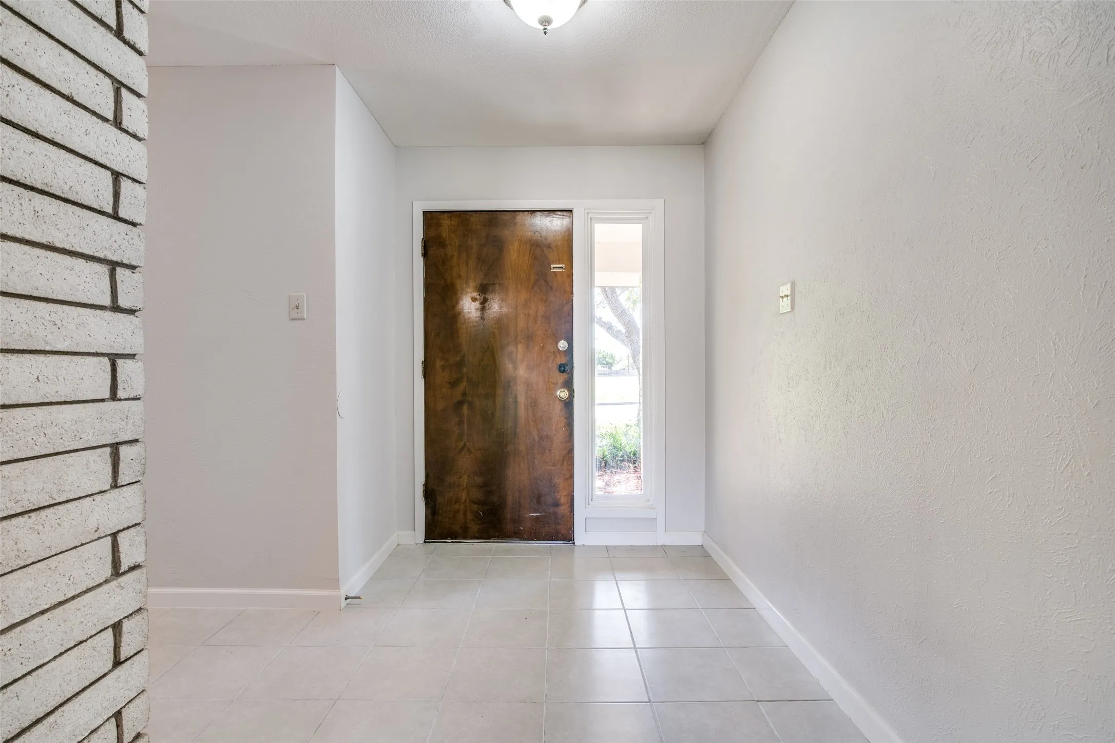 Entryway featuring a dark wood door with a sidelight, light-toned tile flooring, and a white brick architectural element