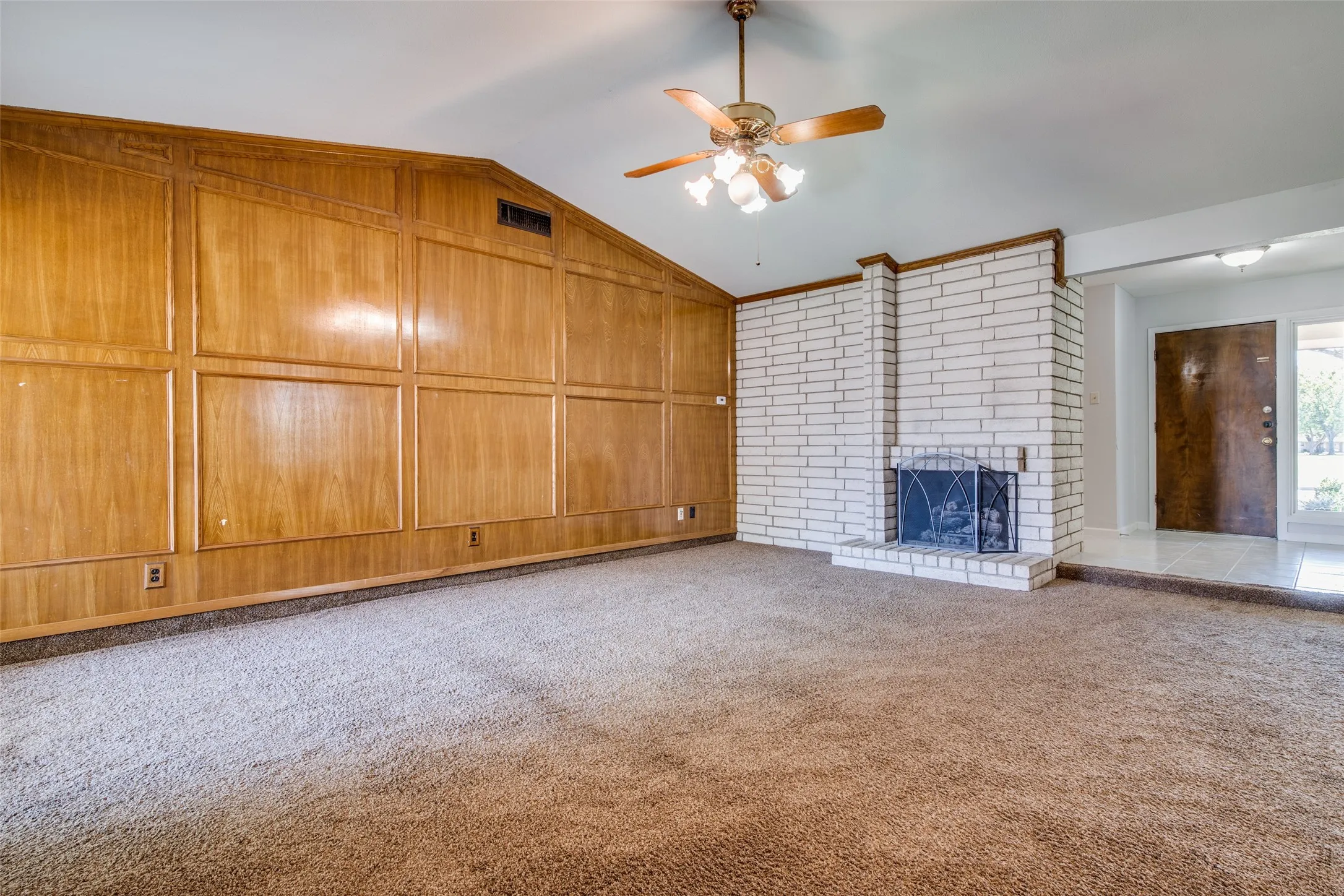 Vaulted ceiling living area featuring a prominent brick fireplace with hearth, wood paneling, and a ceiling fan with integrated lighting