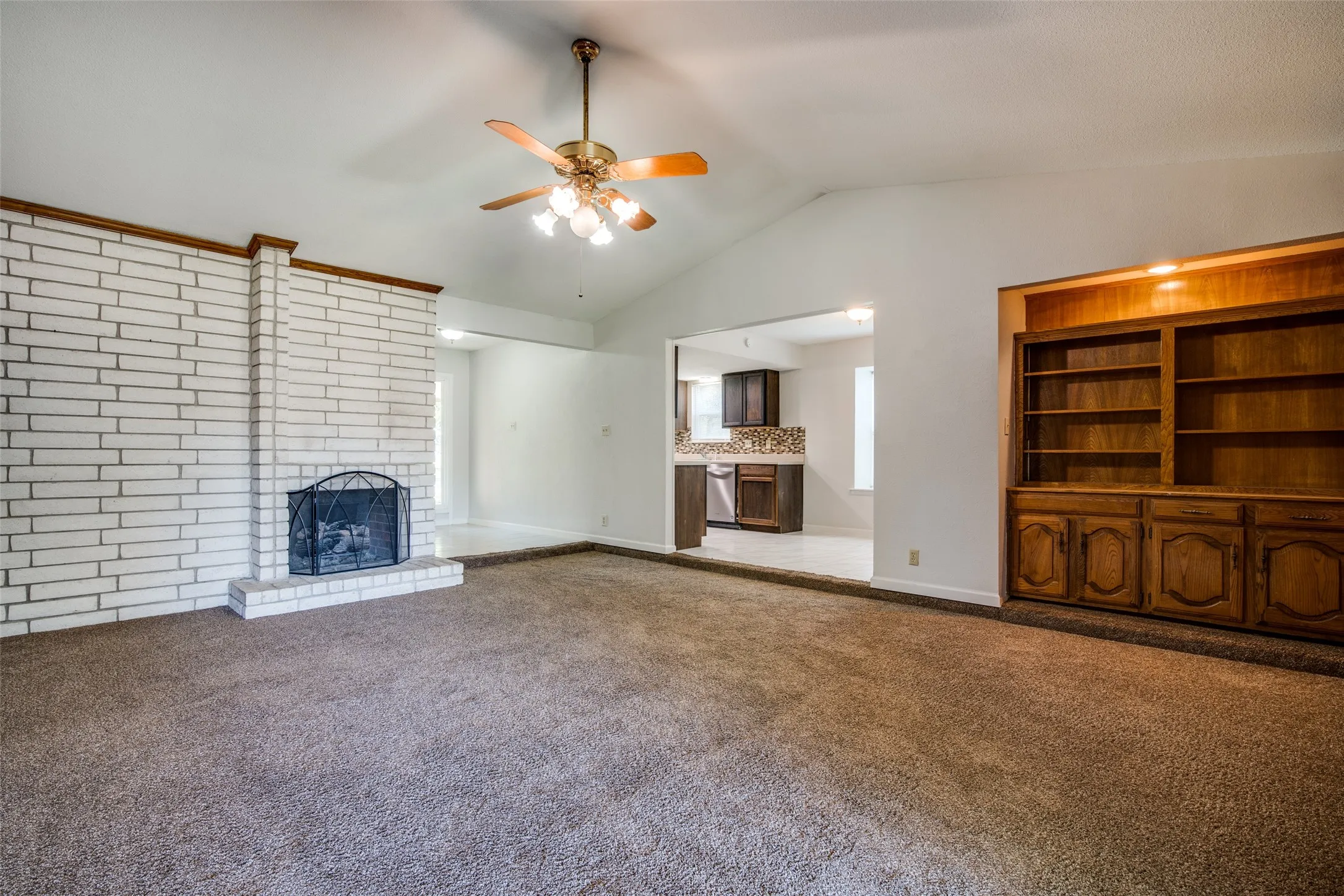Spacious living area featuring a vaulted ceiling with a ceiling fan, a white brick fireplace with a hearth, and built-in wood shelving with lower cabinetry