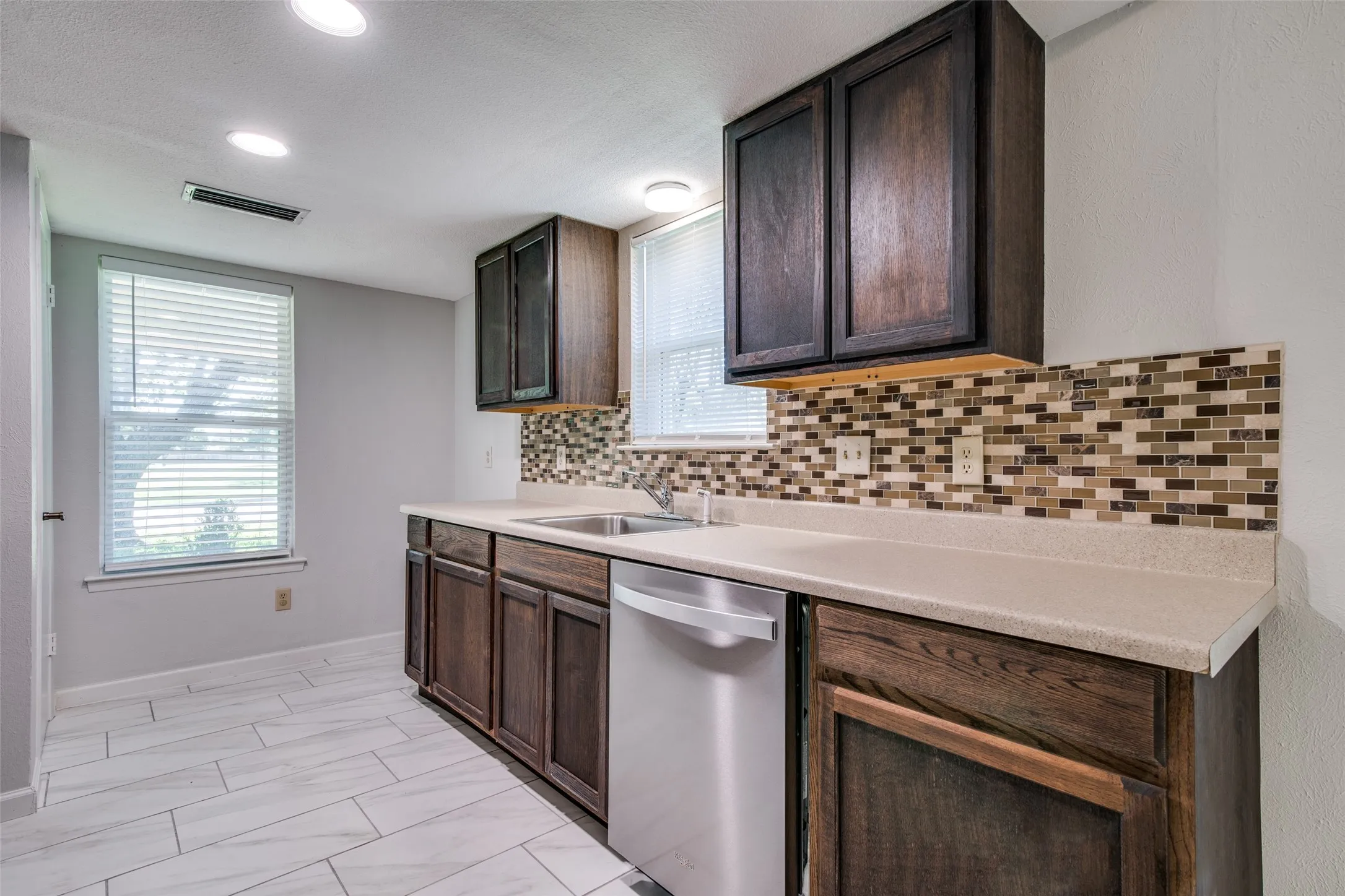 Contemporary kitchen featuring dark wood cabinetry, a mosaic tile backsplash, light-toned countertops, and stainless steel appliances