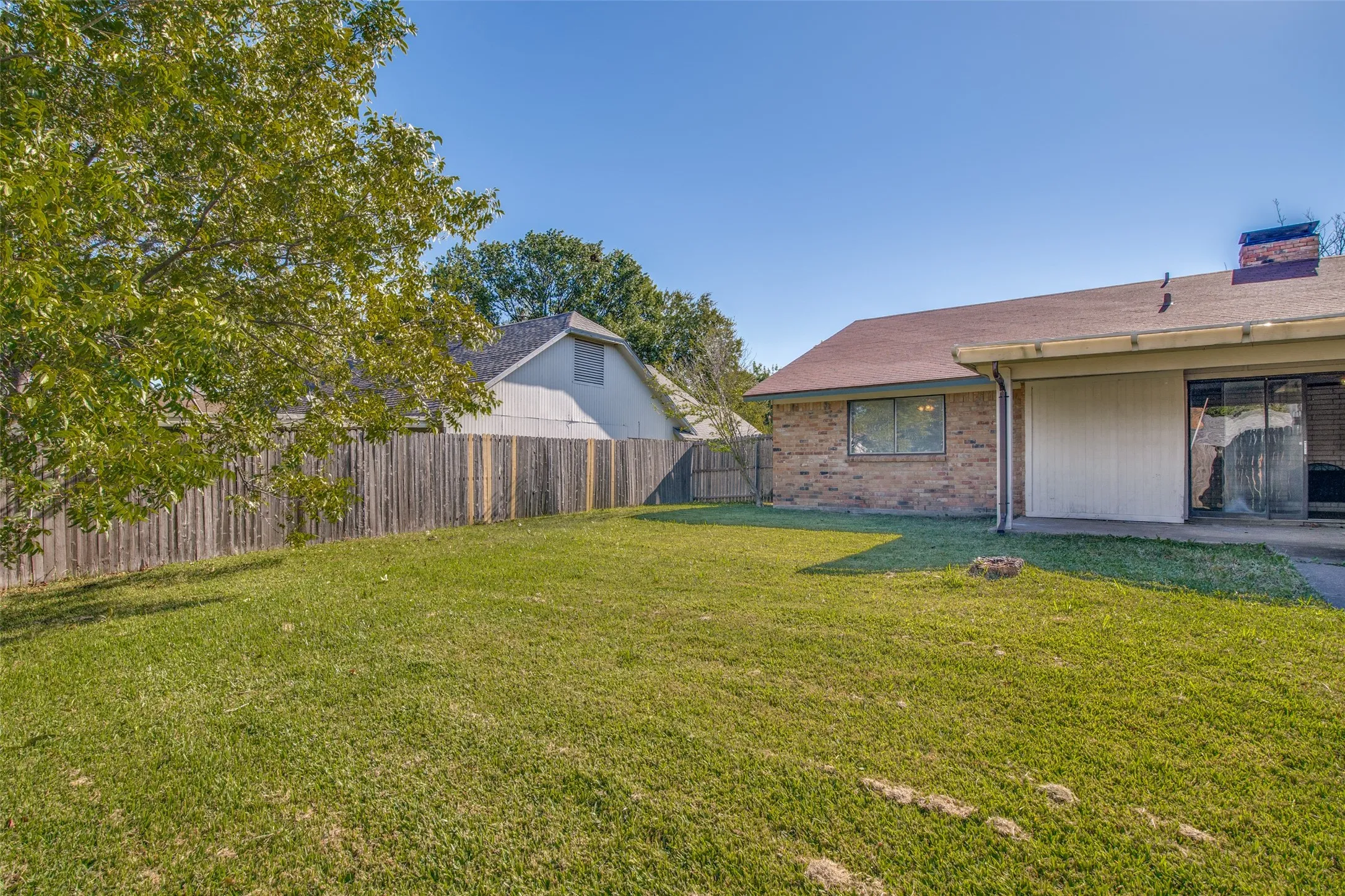 Expansive green lawn with mature perimeter trees, wood privacy fencing, and a brick exterior home featuring a patio area with sliding glass doors