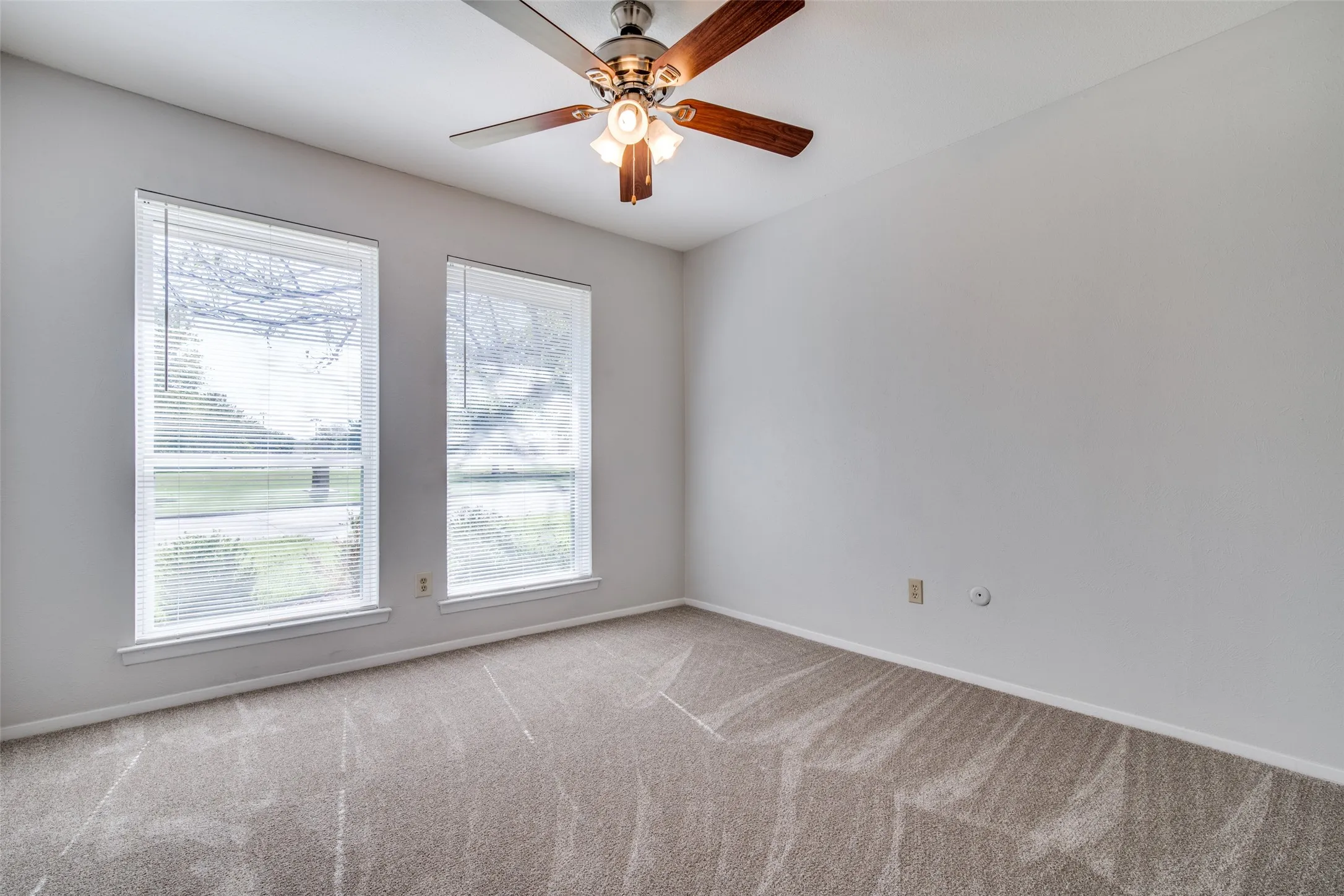 Carpeted room featuring two large windows with blinds, a ceiling fan with light fixture, and light-toned walls