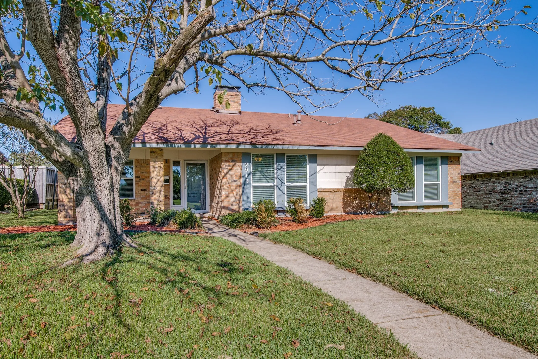 Single-story brick exterior with a red shingle roof, established trees, a concrete pathway, and an attached chimney