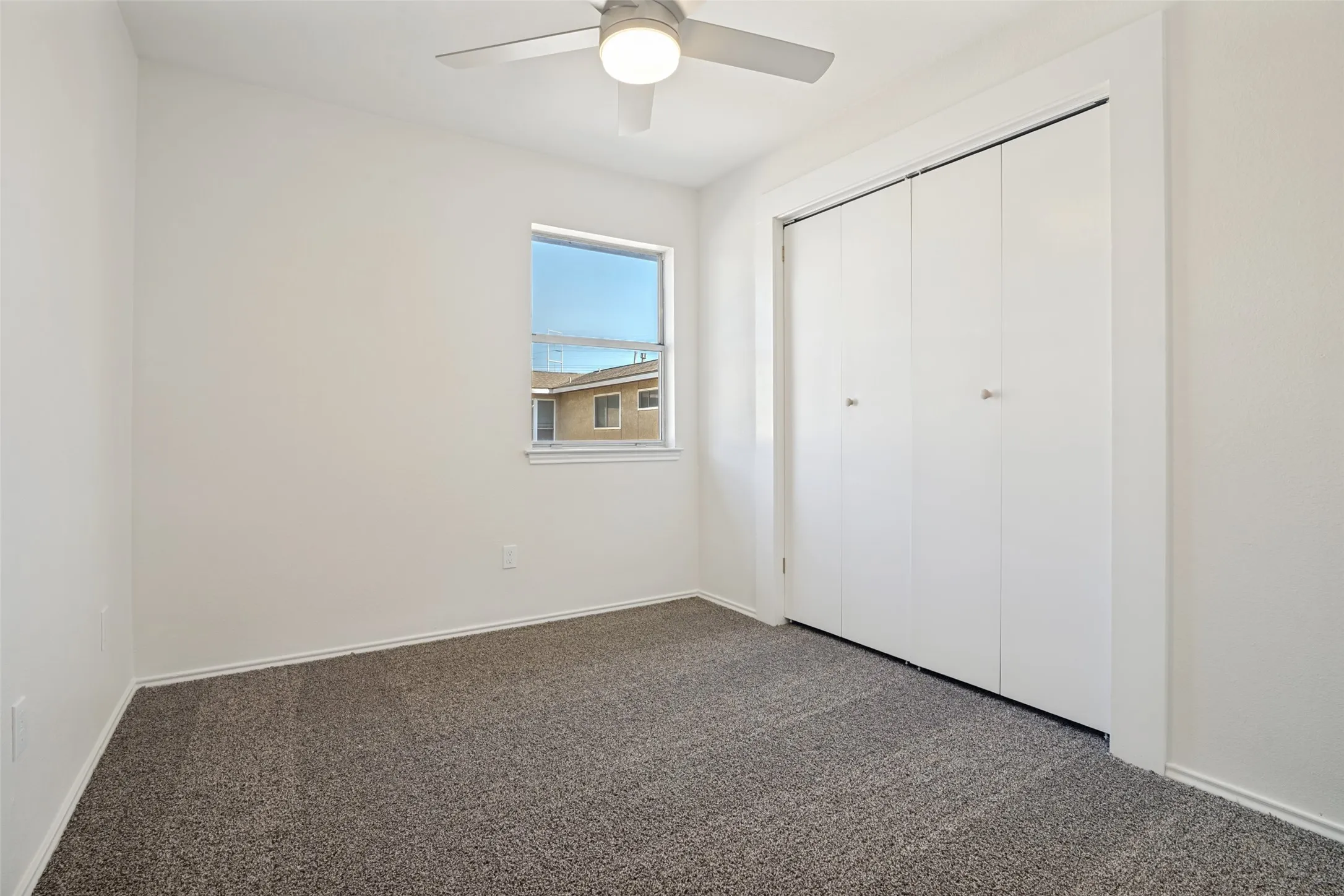Neutral interior space featuring light-colored walls, gray carpet flooring, a window with a white frame, and a ceiling fan with light fixture