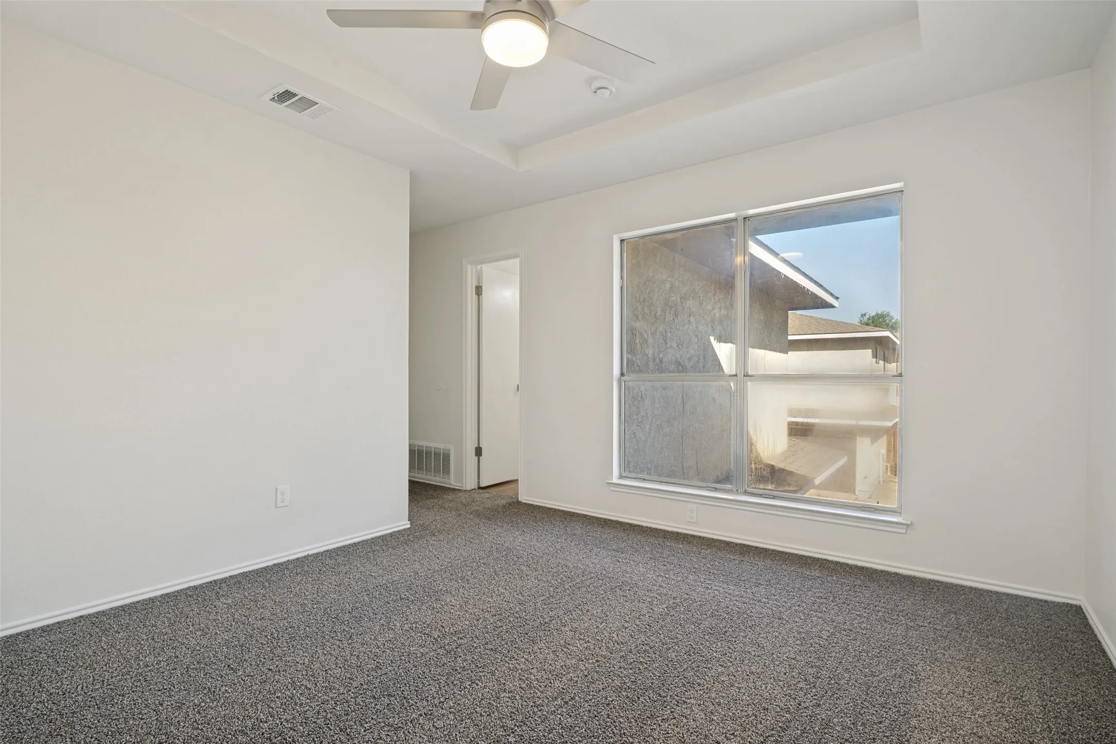 Room featuring a tray ceiling with a flush-mount ceiling fan, a large window, light-colored walls, and gray carpet flooring