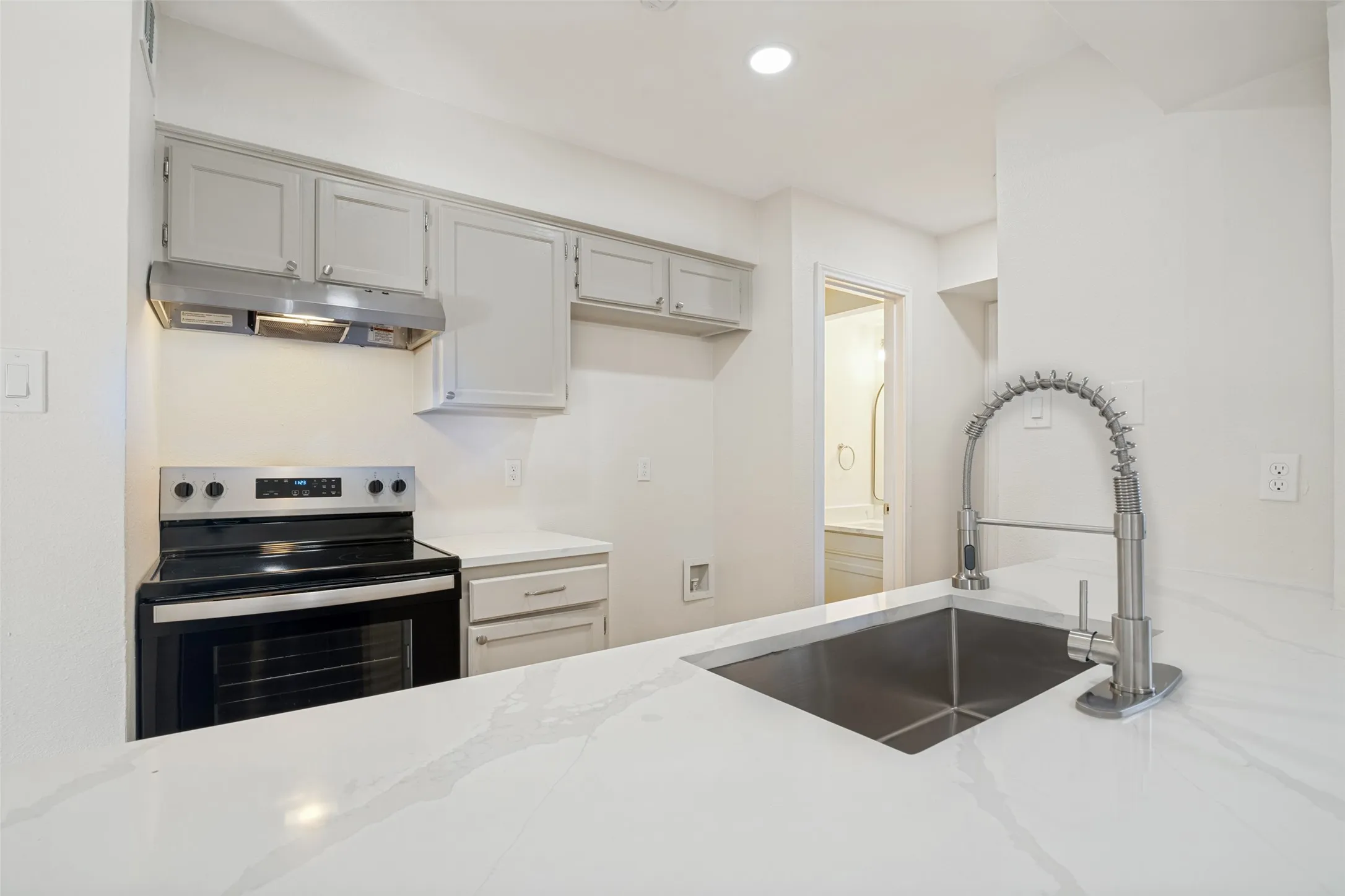Kitchen featuring a stainless steel undermount sink with a gooseneck pull-down faucet, white stone-finish countertops, light gray cabinetry, and a black electric range with an overhead vent hood