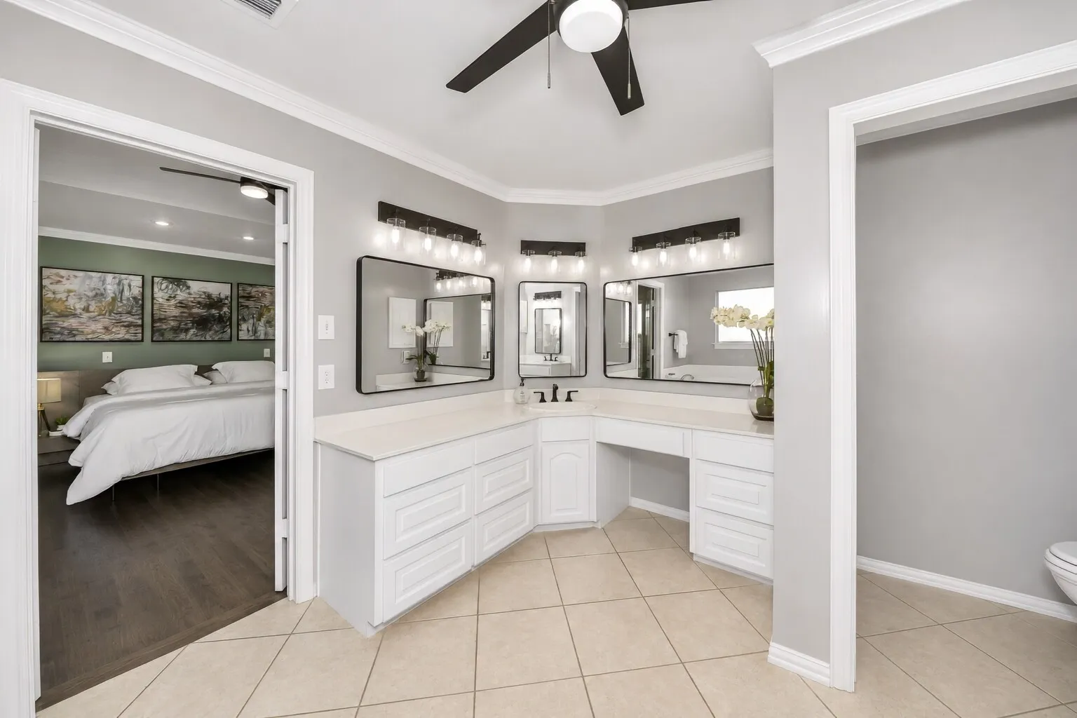 The primary bathroom features a corner three-station vanity with space for a chair, designed for comfortable everyday routines. Framed mirrors and soft, flattering lighting create a bright, modern look that keeps the space feeling calm and functional. White cabinetry and black accents tie everything together, giving the room a timeless style that’s easy to live with.