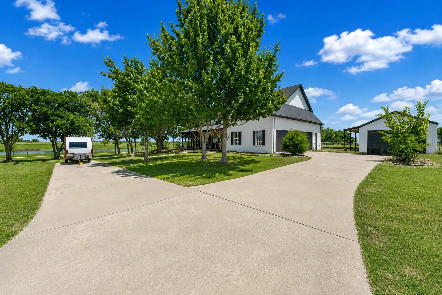 The drive opens into a wide, simple approach, with the farmhouse centered under tall shade trees and clean black-and-white lines. The land stretches out on both sides, offering easy space for boat or RV parking without crowding the view. It’s an open, practical layout that fits the way people actually live — calm, organized, and ready for whatever comes home.