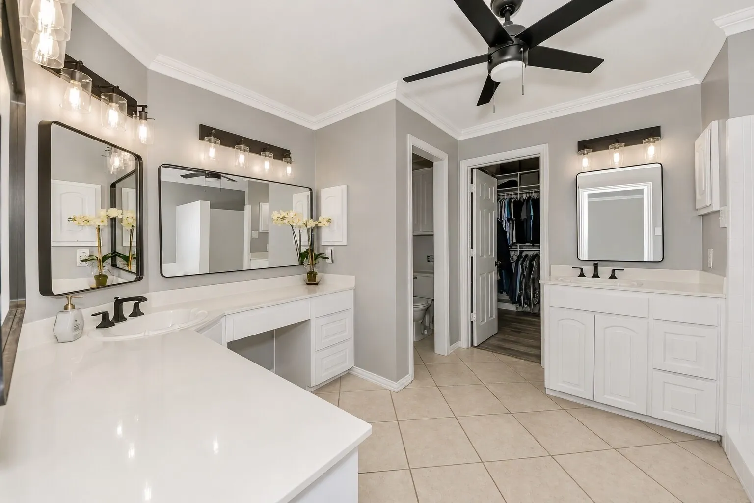 This angle shows the primary bathroom’s clean, modern layout with dual vanities and a dedicated three-station vanity on the left — complete with space for a chair, making morning routines feel relaxed and intentional. The finishes stay crisp and timeless with white cabinetry, black fixtures, and soft natural light. Just beyond the doorway, the custom walk-in closet offers generous storage and an easy flow between getting ready and winding down. It’s a bright, functional space designed to feel calm and effortless.