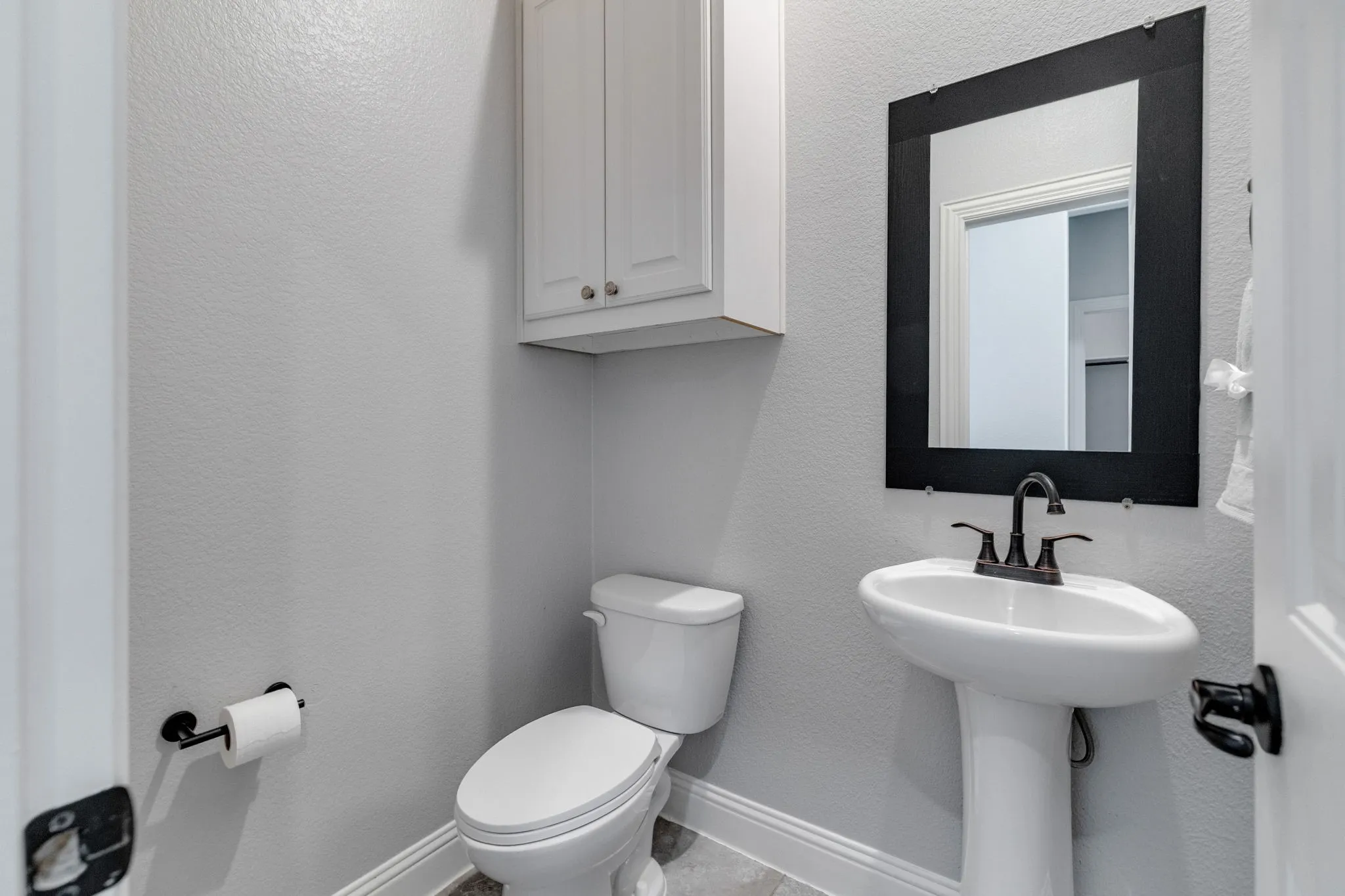 Powder room featuring a pedestal sink with a dark bronze faucet, a framed wall mirror, and white upper cabinetry