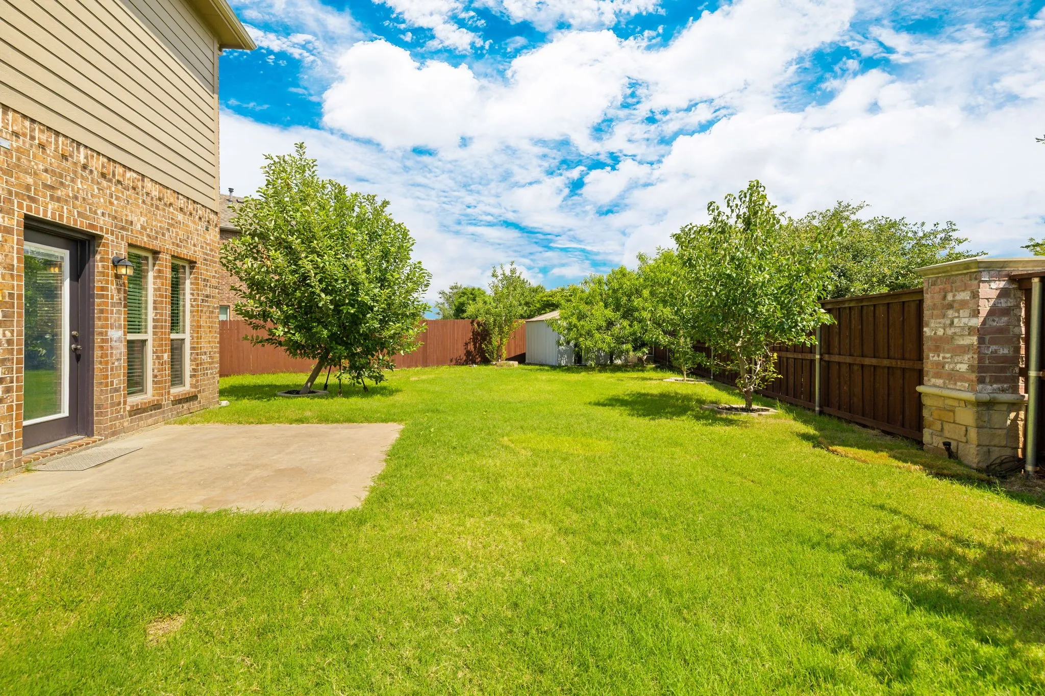 Spacious backyard featuring a large concrete patio, mature trees, wood privacy fencing with brick columns, and a storage shed