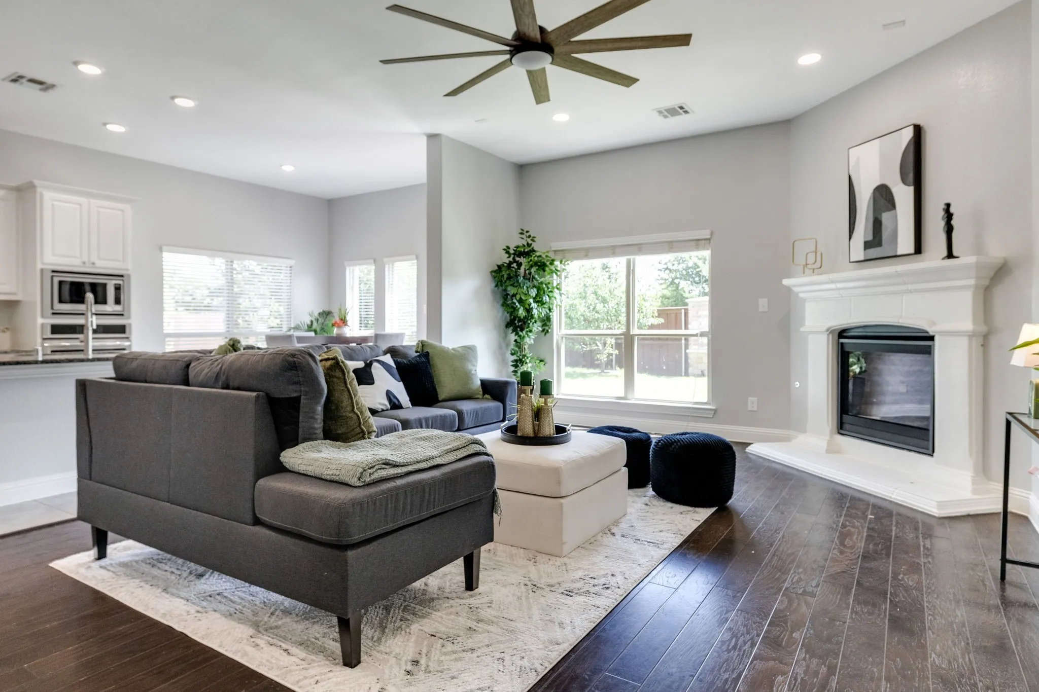 Expansive living area featuring rich wood-finish flooring, a white-mantled fireplace, and a contemporary ceiling fan