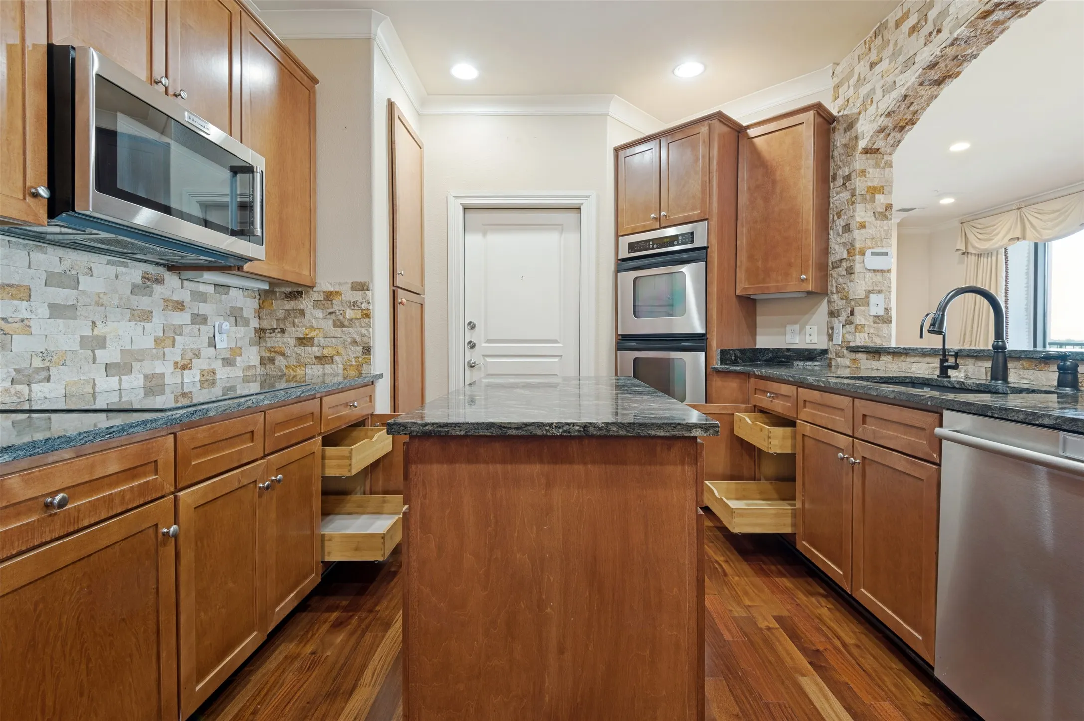 Gourmet kitchen with rich wood cabinetry with pull-out storage drawers, stone tile backsplash, built-in microwave, stainless steel appliances, high-end granite countertops, stone tile backsplash, and a portable central island