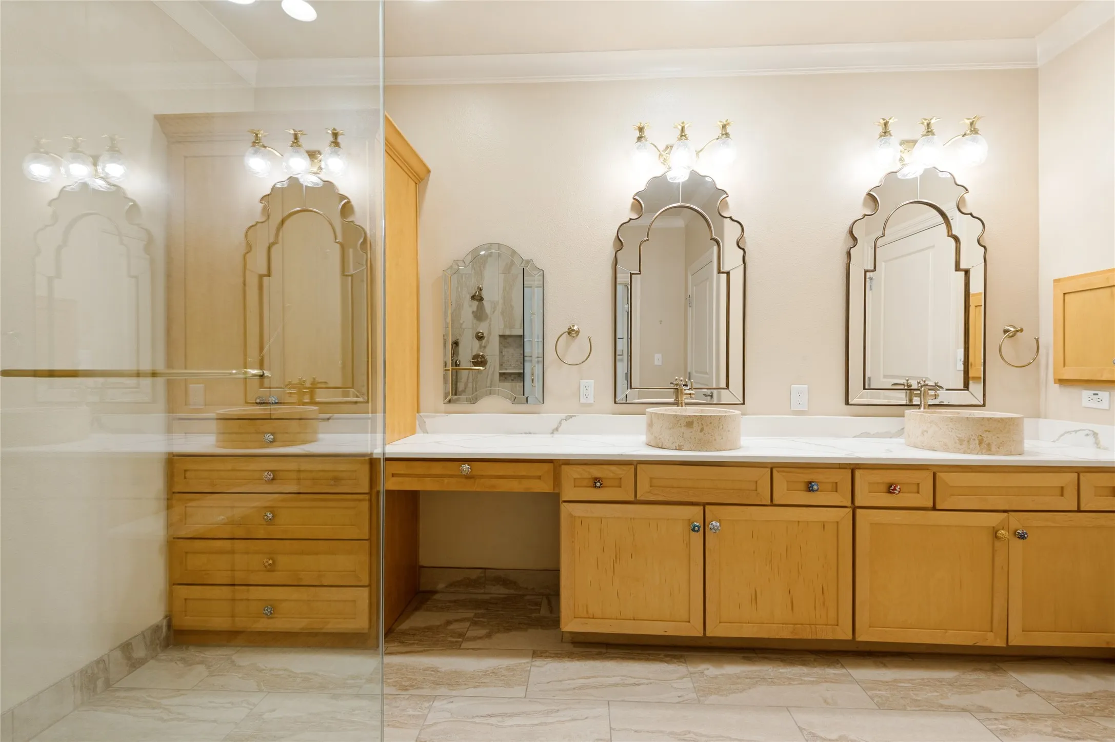 Dual stone vessel sinks set into a light-colored countertop and a massive walk-in shower with built-in shower seat. Vanity feature an abundance of storage, Venetian mirrors, marble countertops and designer lighting