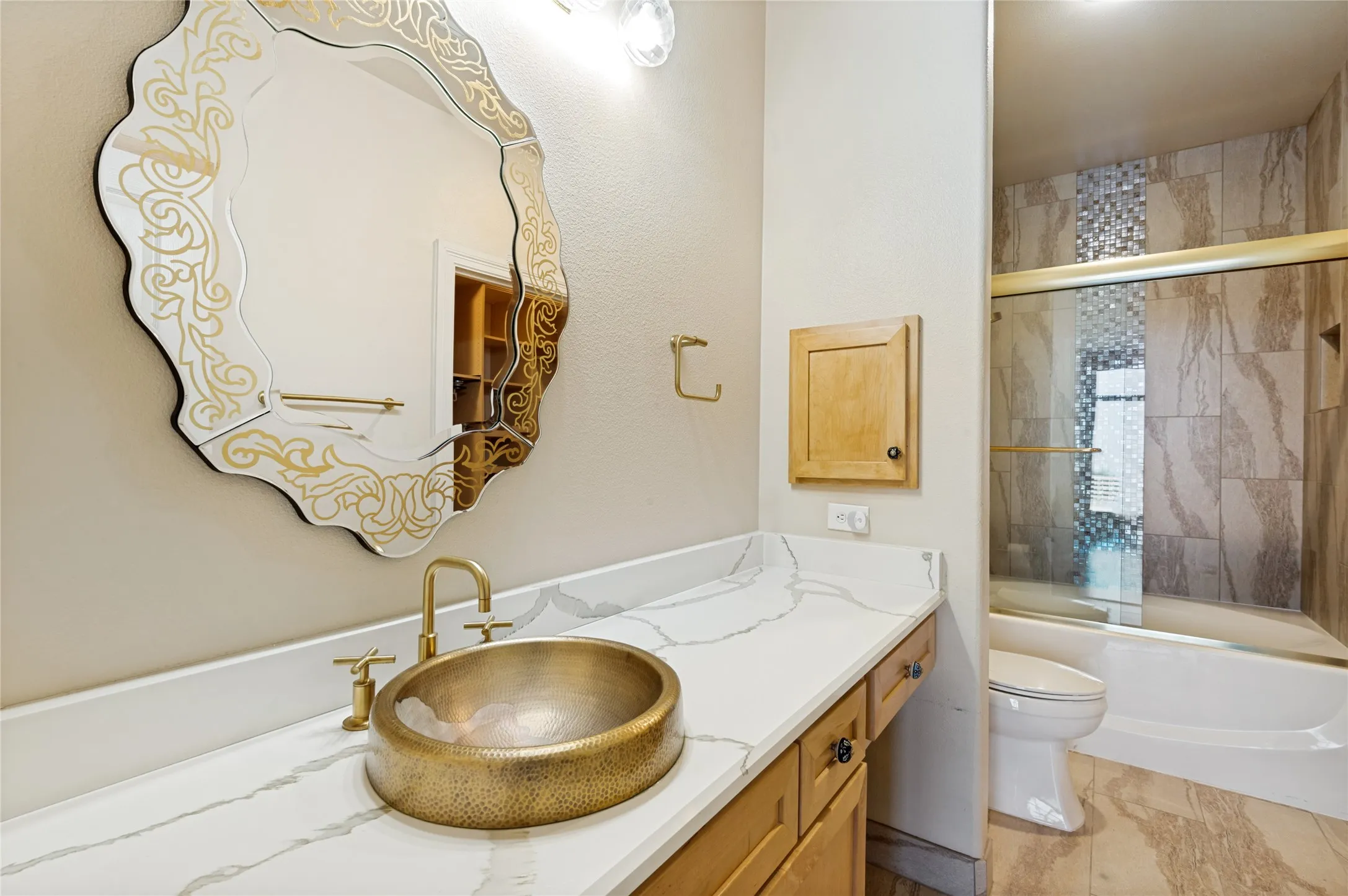 Bathroom vanity featuring a vessel sink with a hammered brass finish, a white countertop with grey veining, and natural wood-finish cabinetry