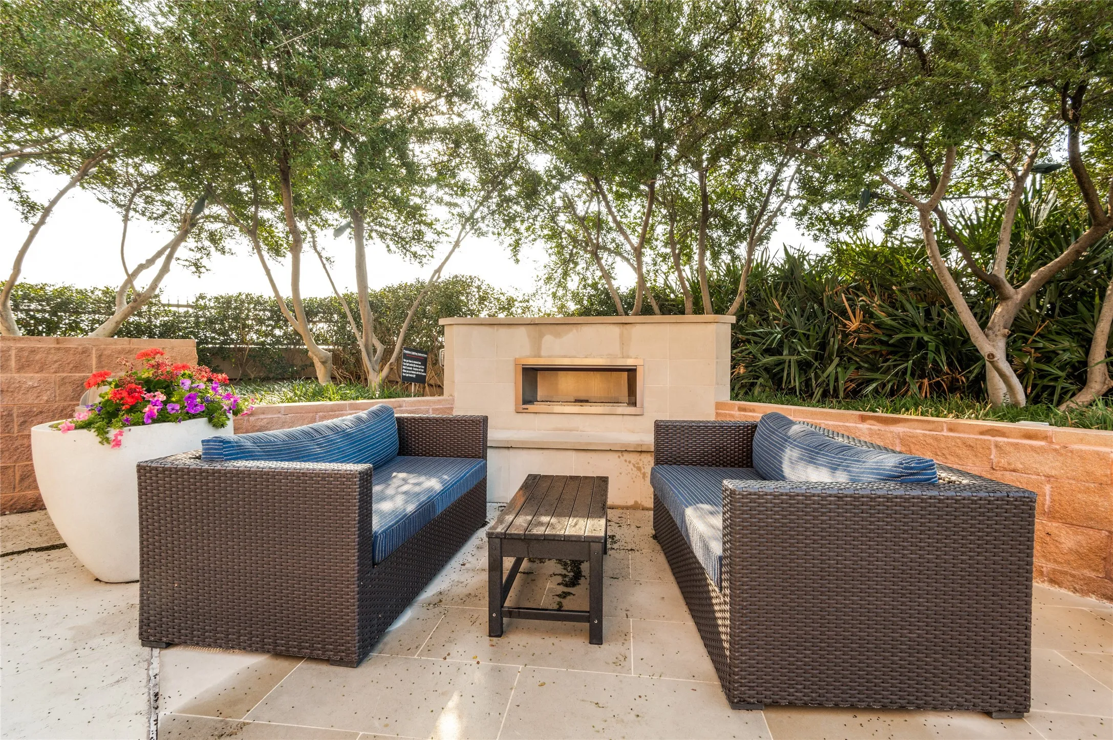 Outdoor patio featuring a built-in fireplace, natural stone block walls, mature trees, and light-colored paver flooring
