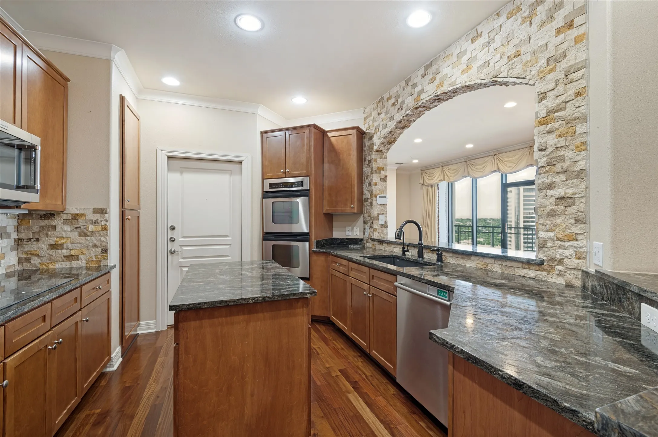 Kitchen featuring wood-finish flooring, stone countertops, integrated stainless steel appliances, and an arched pass-through window with a stone-clad surround