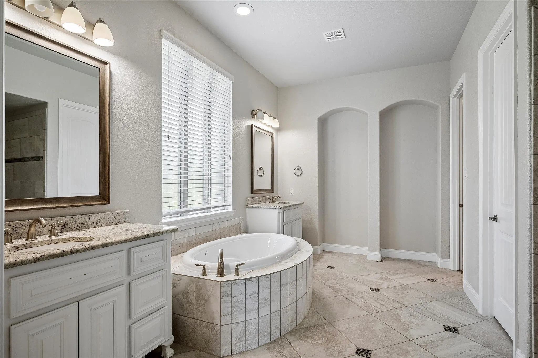 Spacious bathroom featuring a built-in soaking tub with tiled surround, dual vanity areas with granite countertops and undermount sinks, tile flooring, and arched wall niches
