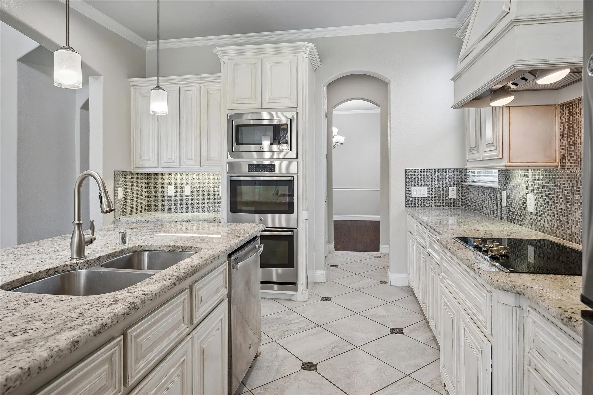 Spacious kitchen featuring an island with a double stainless steel sink, granite countertops, white cabinetry, built-in stainless steel appliances, and a mosaic tile backsplash