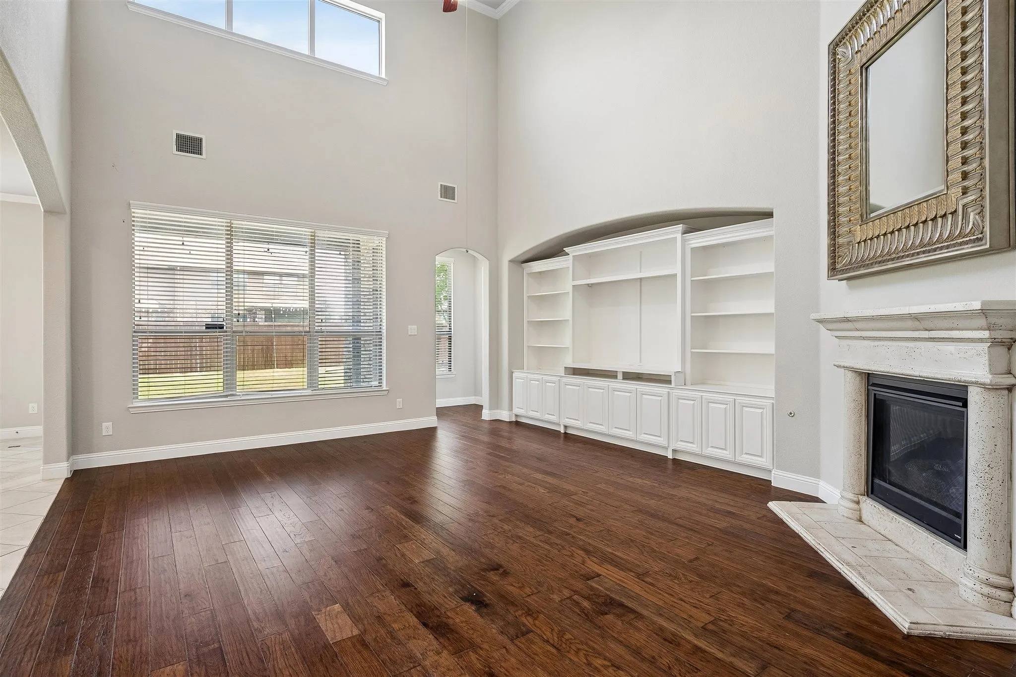 Spacious living area featuring high ceilings, wood-finish flooring, a stone fireplace with hearth, and extensive built-in cabinetry with shelving
