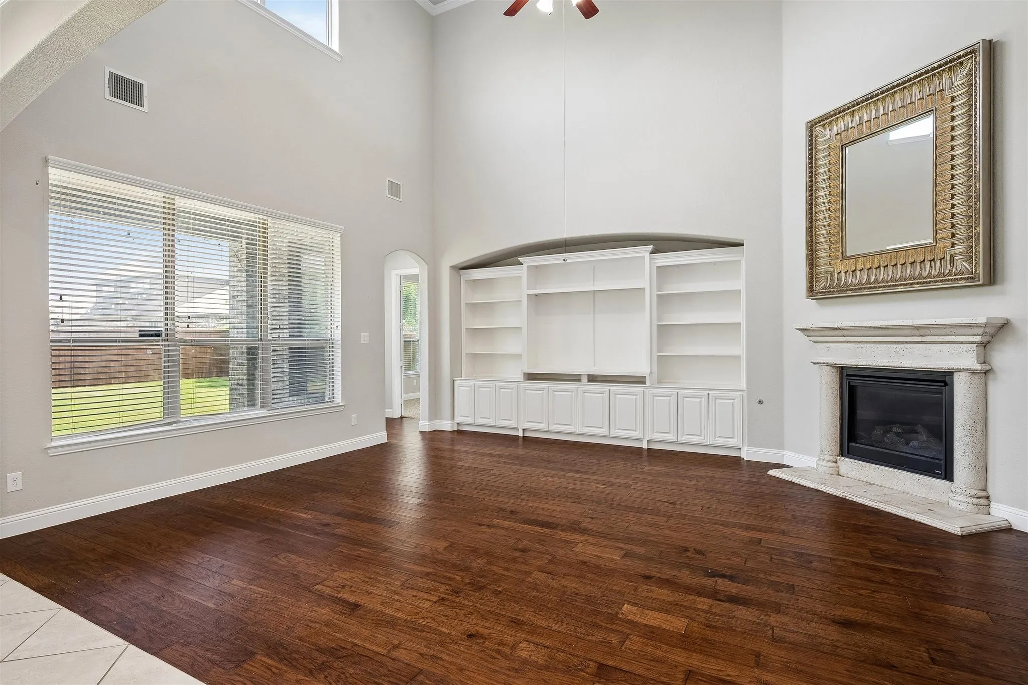 Spacious living area featuring high ceilings, wood-finish flooring, a built-in media center with shelving, a fireplace with a stone surround, and large windows overlooking the backyard