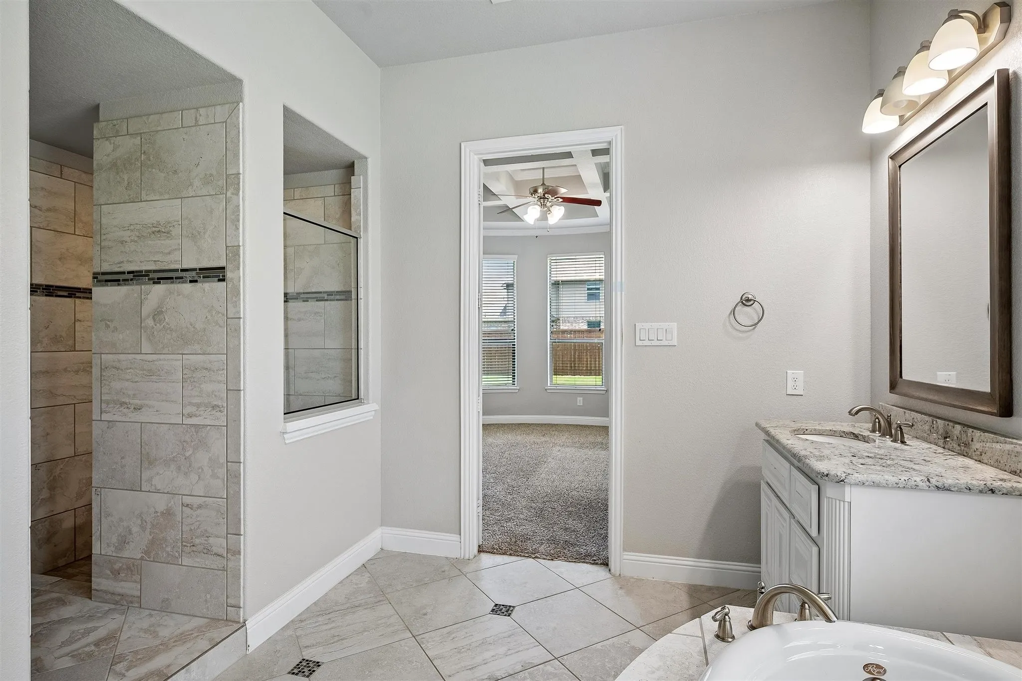 Bathroom featuring tile flooring with decorative inserts, a built-in shower with stone-look tile, a separate built-in soaking tub, a vanity with a light-colored countertop and undermount sink, and a framed mirror