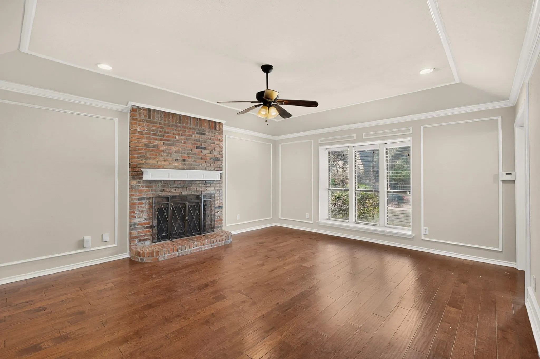 Spacious living area featuring a prominent brick fireplace with a white mantel, wood-finish flooring, and a tray ceiling with recessed lighting