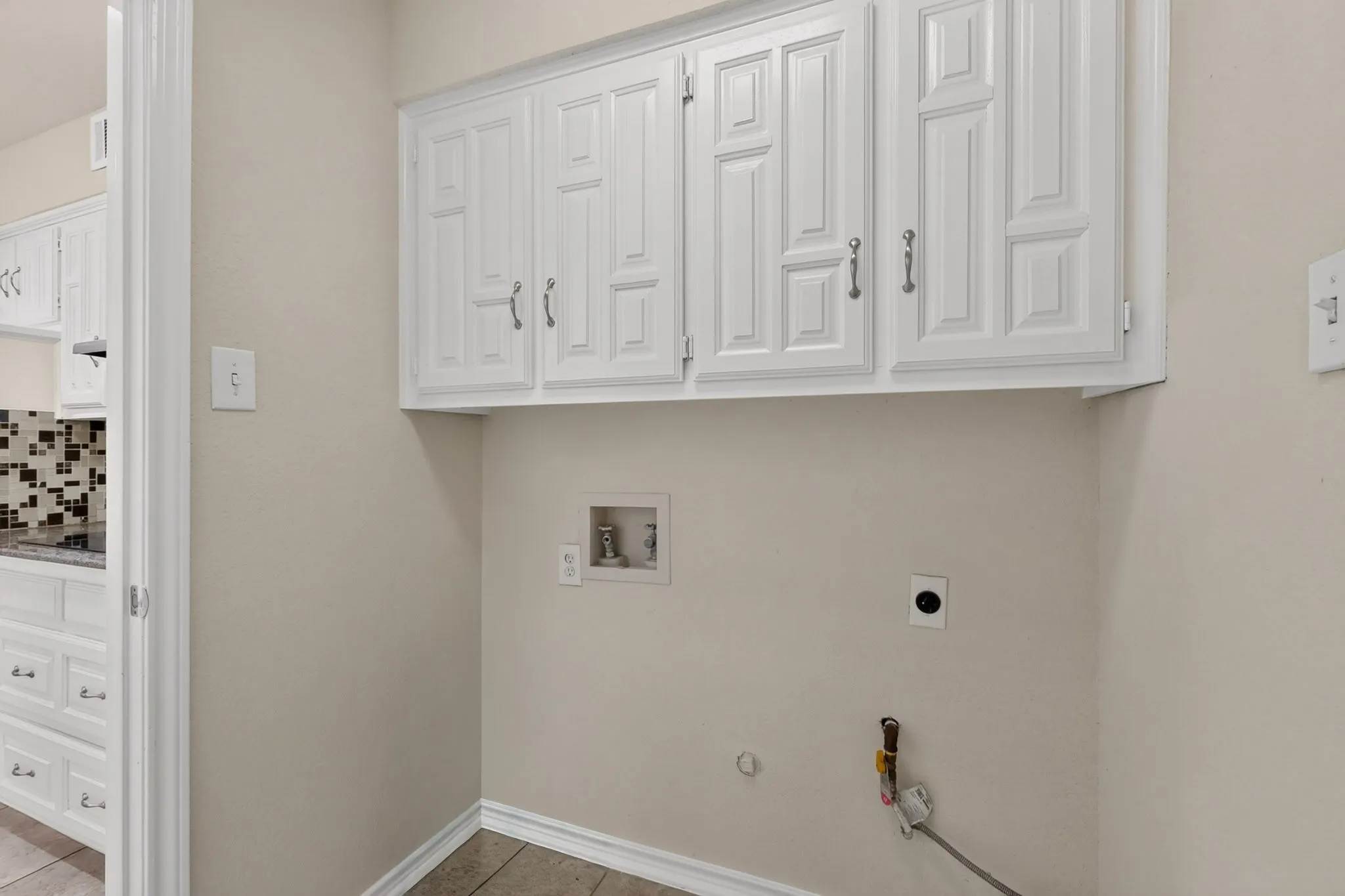 Utility area featuring white paneled upper cabinetry with chrome hardware, neutral wall paint, and tiled flooring
