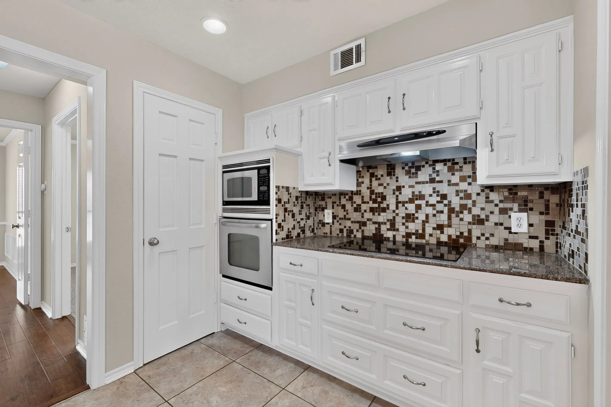 Kitchen featuring white cabinetry with brushed nickel hardware, a patterned mosaic tile backsplash, built-in stainless steel microwave and oven, a built-in cooktop, and stone-finish countertops