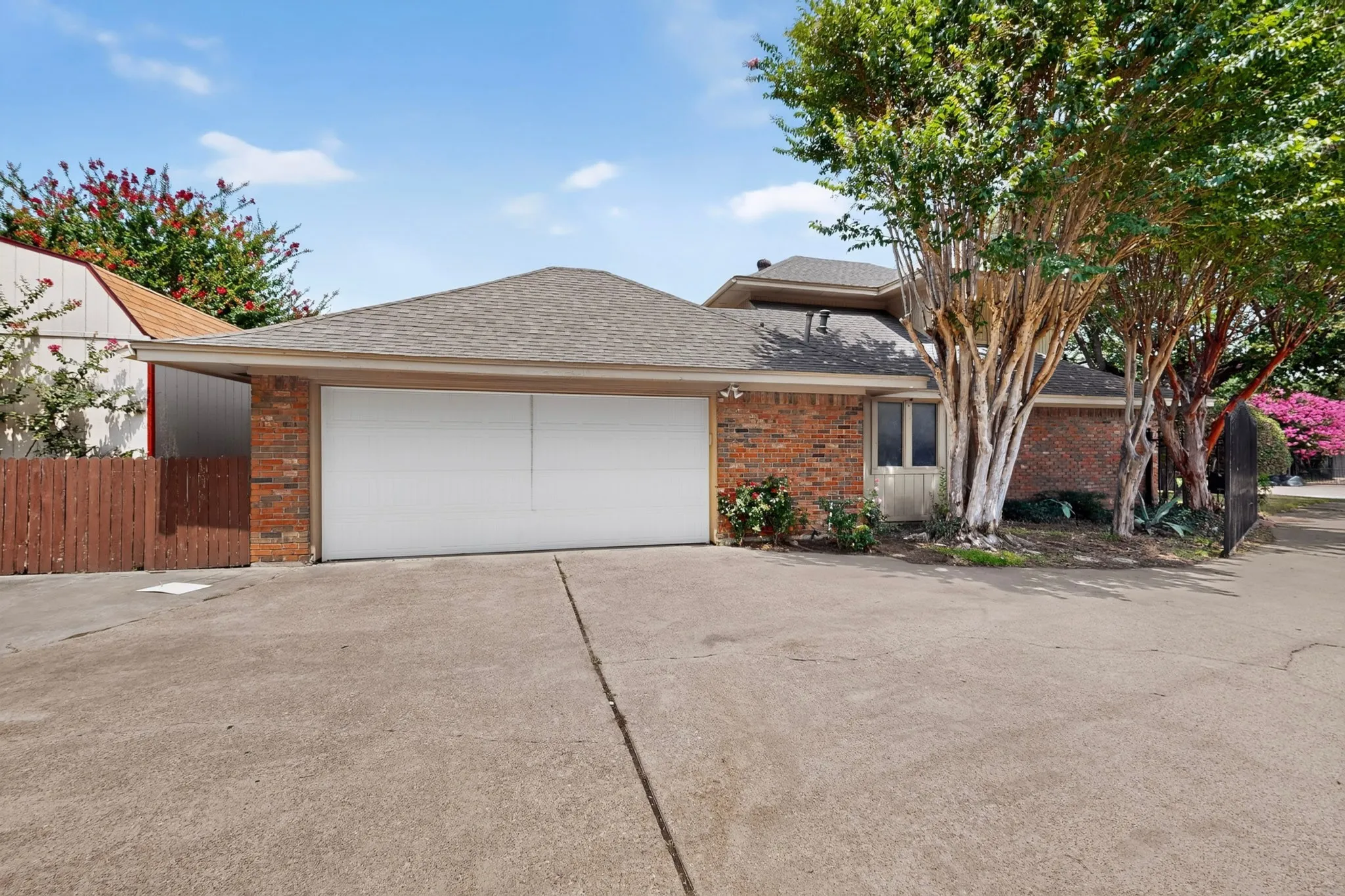 Brick exterior featuring a two-car garage with a concrete driveway, a gabled roofline, and mature trees framing the entrance