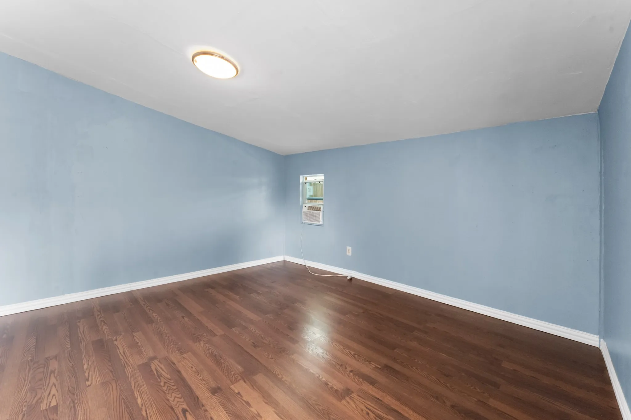 Hardwood flooring, light blue wall paint, white trim, window-mounted air conditioning unit, and flush-mount ceiling light