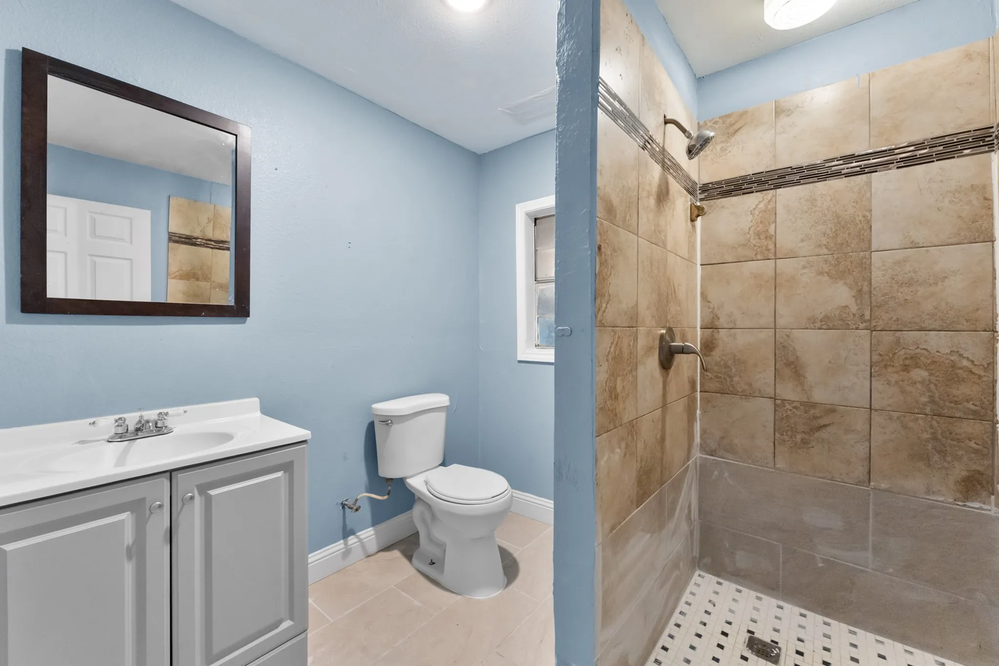 Walk-in shower with tiled surround and decorative accent band, light-colored flooring, vanity with integrated sink, and framed mirror