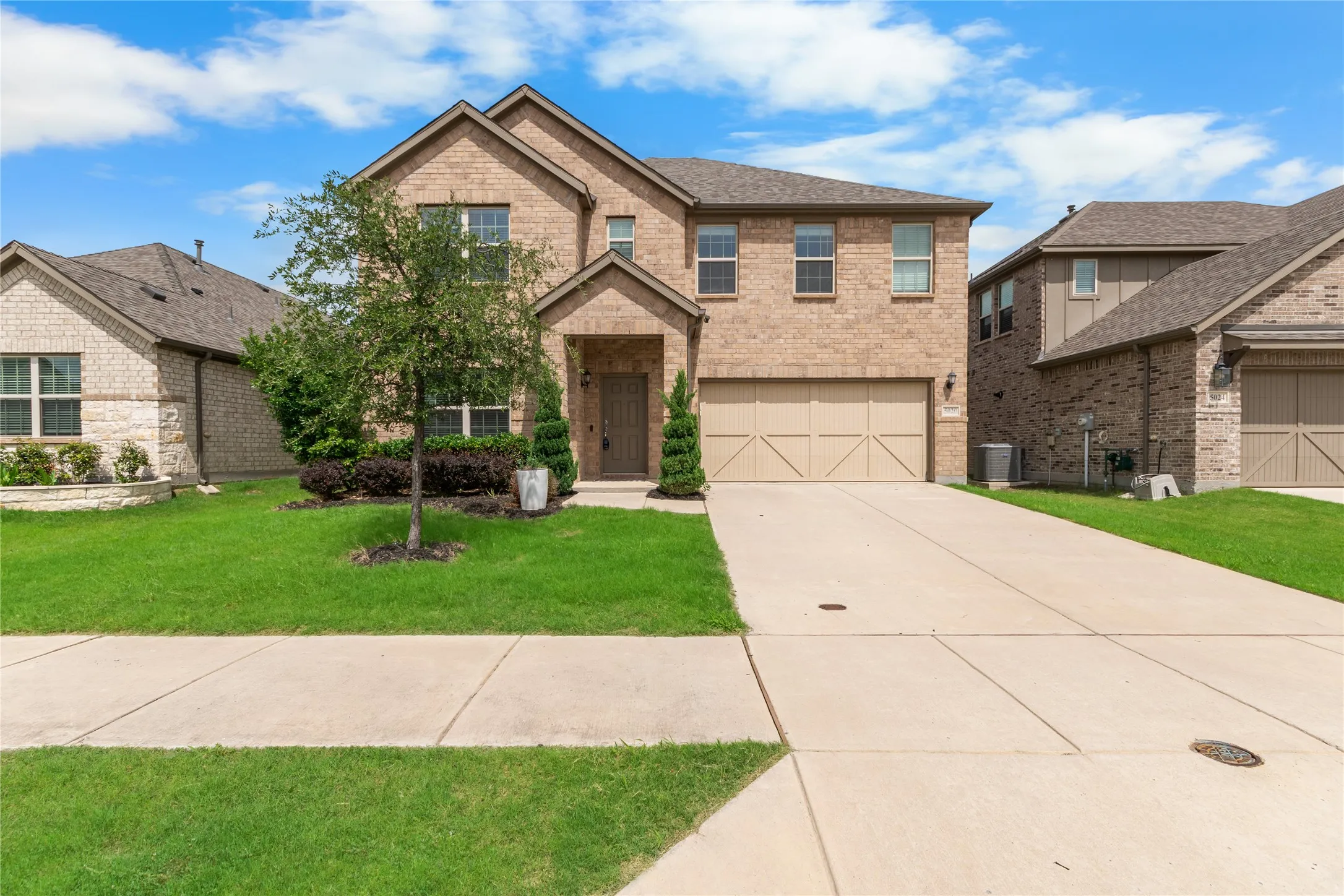 Brick veneer two-story residence featuring an attached two-car garage with a panel door, a concrete driveway, and a front yard with mature landscaping