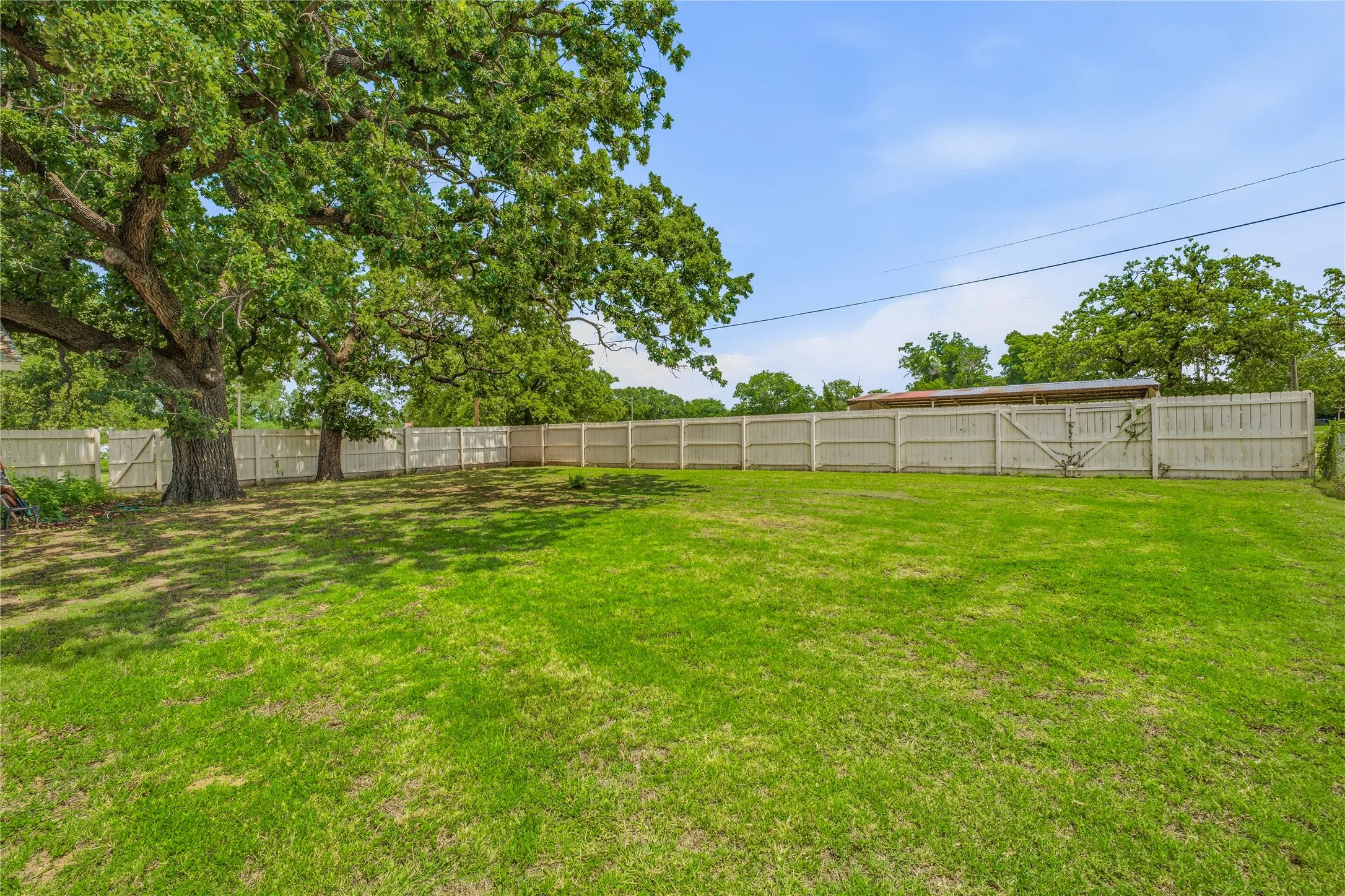 Spacious grassy yard featuring mature trees and a perimeter privacy fence with a gate