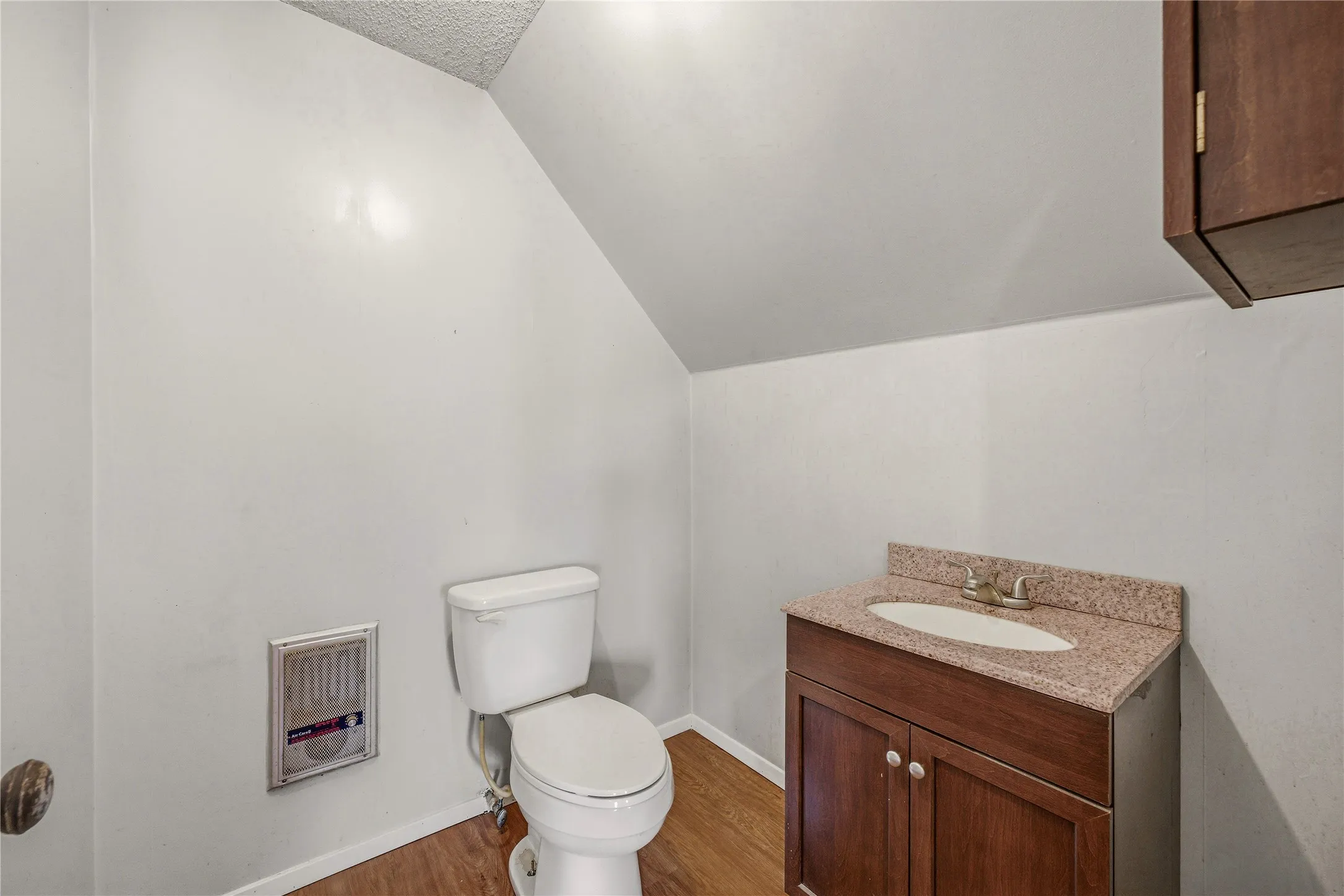 Compact bathroom featuring wood-finish flooring, a white toilet, and a vanity with a light-toned stone-finish countertop and dark wood cabinetry