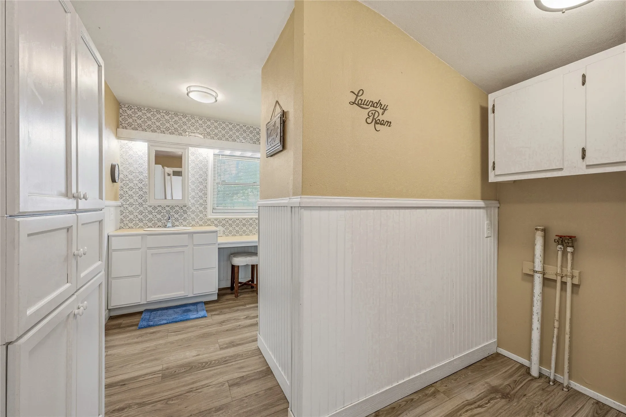 Laundry area featuring wood-finish flooring, white beadboard wainscoting, built-in white cabinetry, a vanity with a sink, and a window