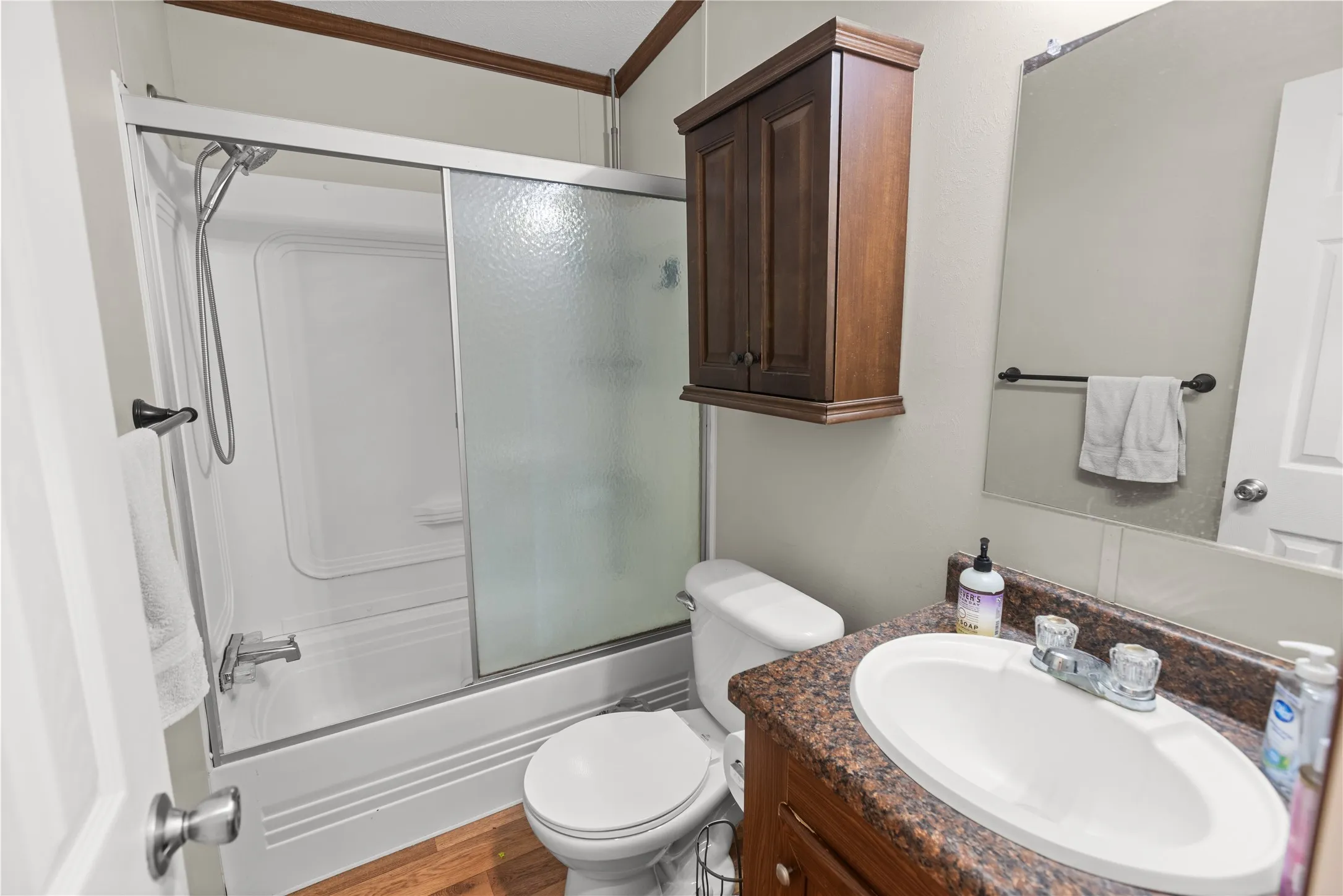 Bathroom featuring a white tub/shower combination with frosted glass sliding doors, a vanity with a dark speckled countertop and integrated sink, wood-finish flooring, and a wall-mounted wood cabinet