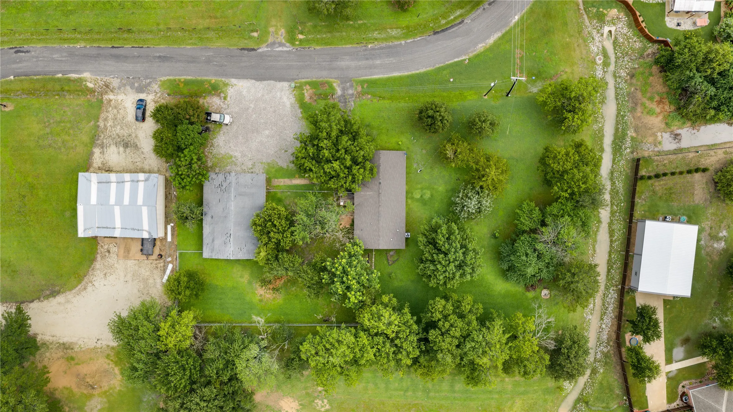 Expansive property featuring a main residence, an additional structure with a metal roof, a gravel driveway, and a large grassy area with mature trees