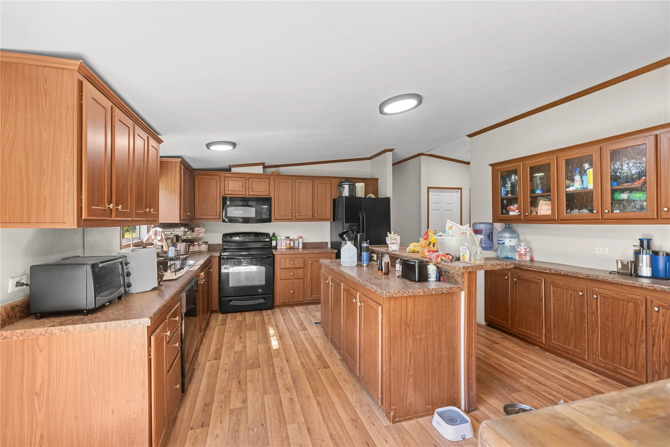 Expansive kitchen featuring wood-finish cabinetry, black appliances, a central island, wood-finish flooring, and ample counter space