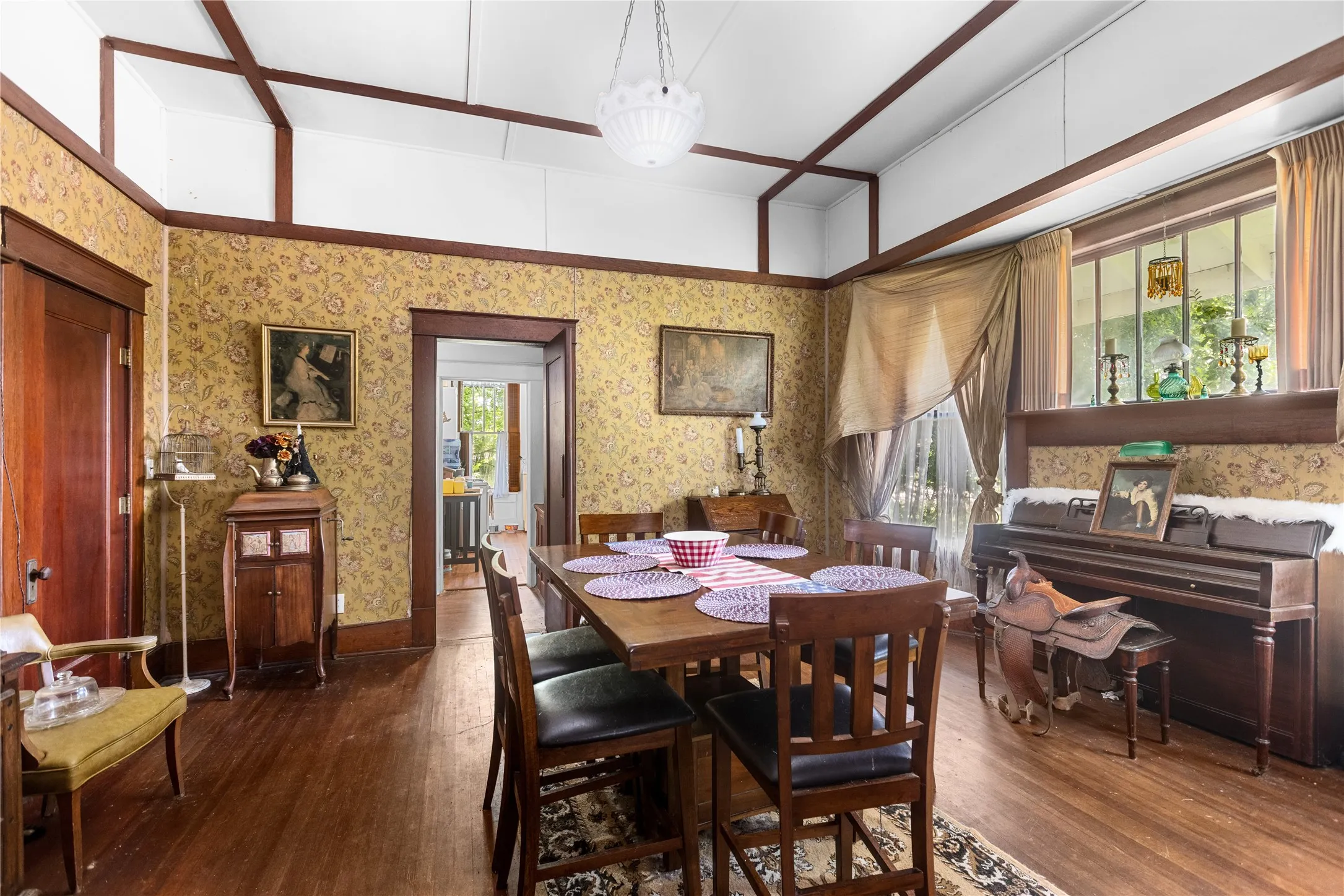 Dining area featuring wood-finish flooring, patterned wallpaper, exposed ceiling beams, a central chandelier, and a window with architectural divisions