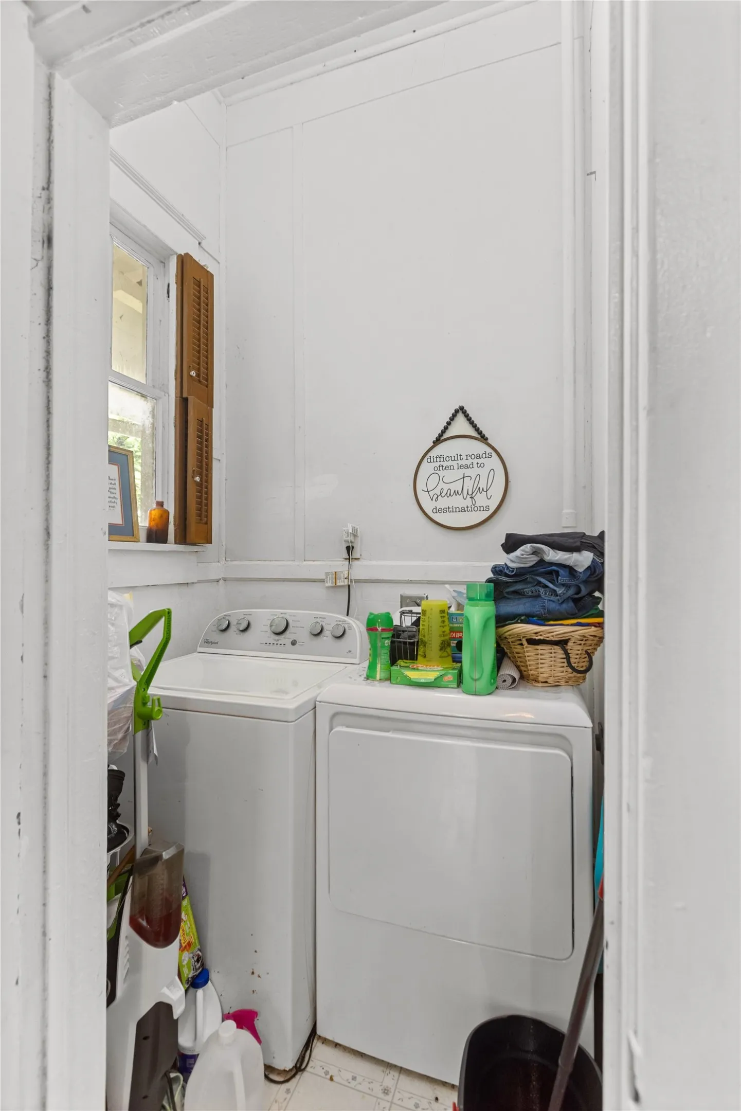 Compact utility space featuring a window with wooden louvered shutters, white wall paneling, and a white tile-finish floor
