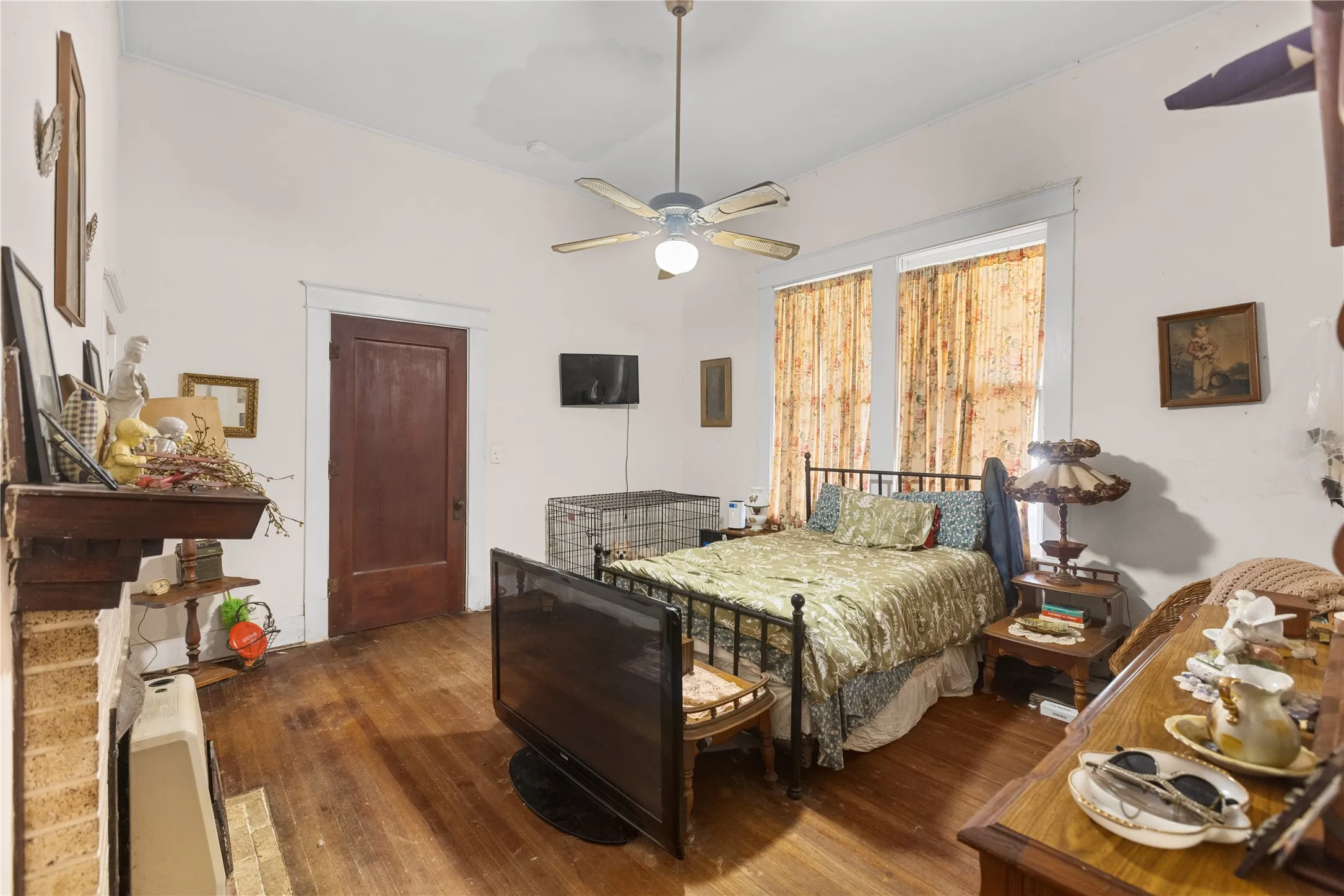 Room featuring wood-finish flooring, two large windows, a ceiling fan, a brown wood door, and a brick fireplace with a wood mantle