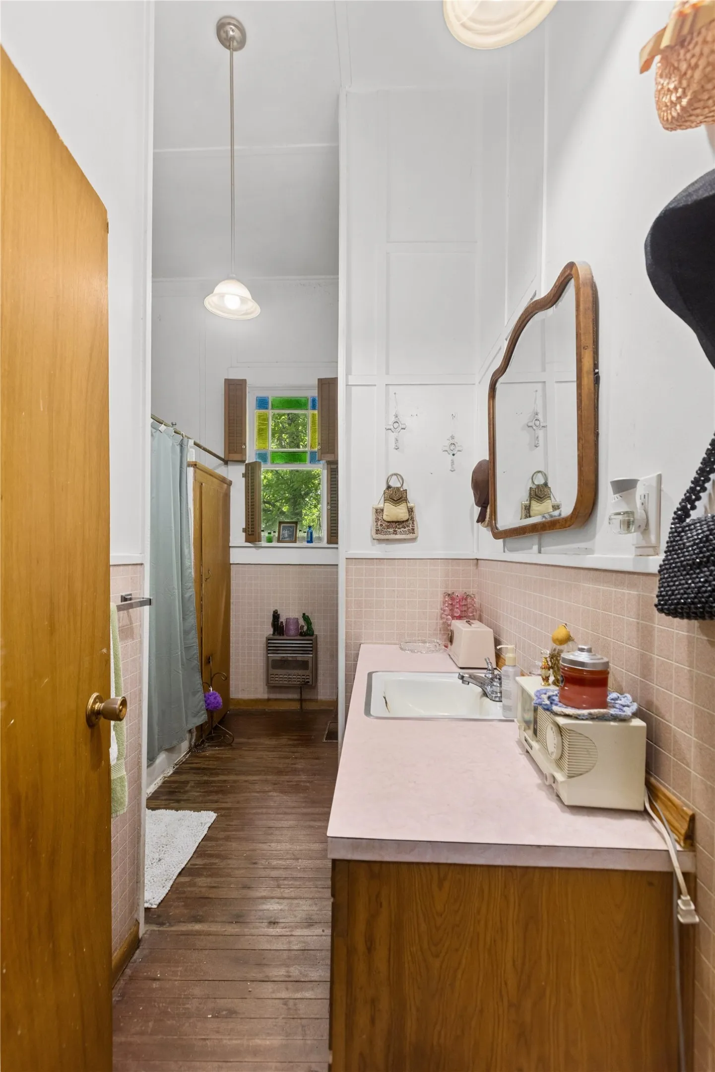 Wood-finish flooring throughout, featuring a vanity with a pink countertop and integrated sink, light pink tiled walls, and a stained glass window