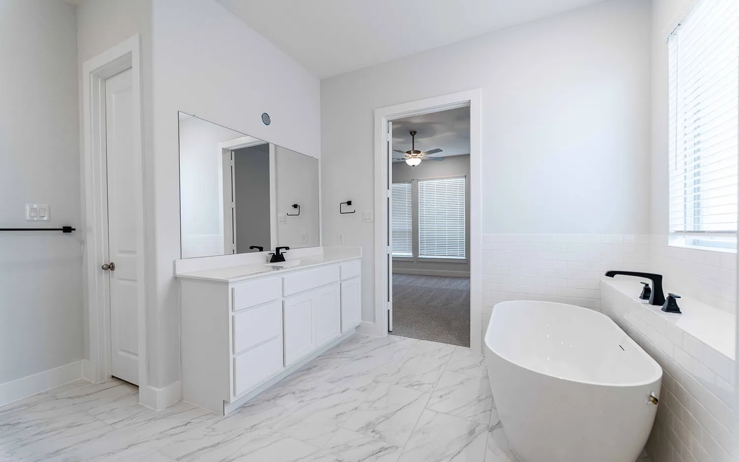 Spacious bathroom featuring a freestanding tub, white vanity with dual sinks, white tile flooring, white horizontal tile accents, and a large mirror