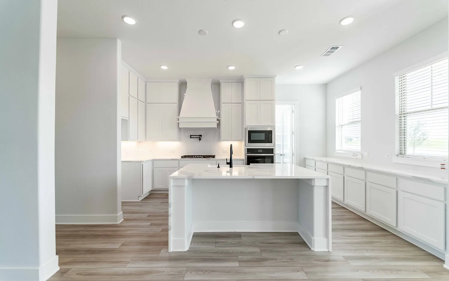 Open-concept kitchen featuring an oversized island with a built-in sink, white shaker cabinetry, wood-finish flooring, recessed lighting, and integrated stainless steel appliances