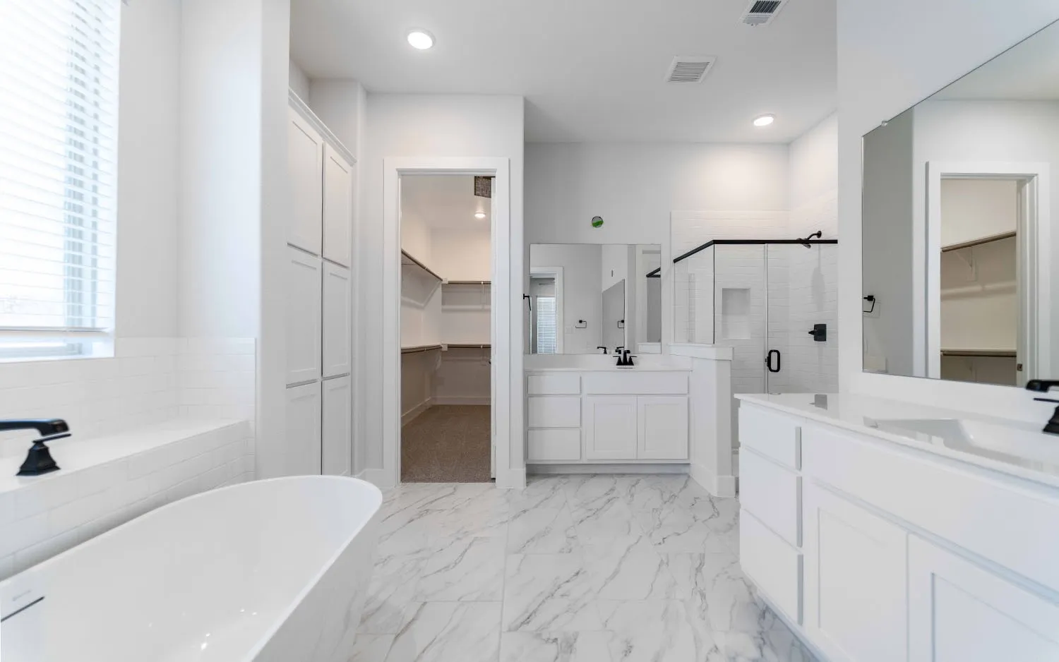 Spacious bathroom featuring a freestanding tub, dual vanities with white cabinetry, a glass-enclosed shower with black trim, and marble-look flooring