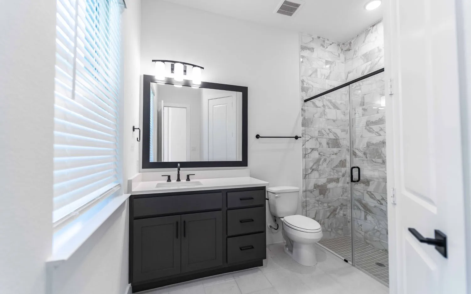 Modern bathroom featuring a dark wood-finish vanity with a white countertop, a rectangular framed mirror with a three-light fixture, a walk-in shower with a glass enclosure and marble-look tile, and light-toned floor tiling