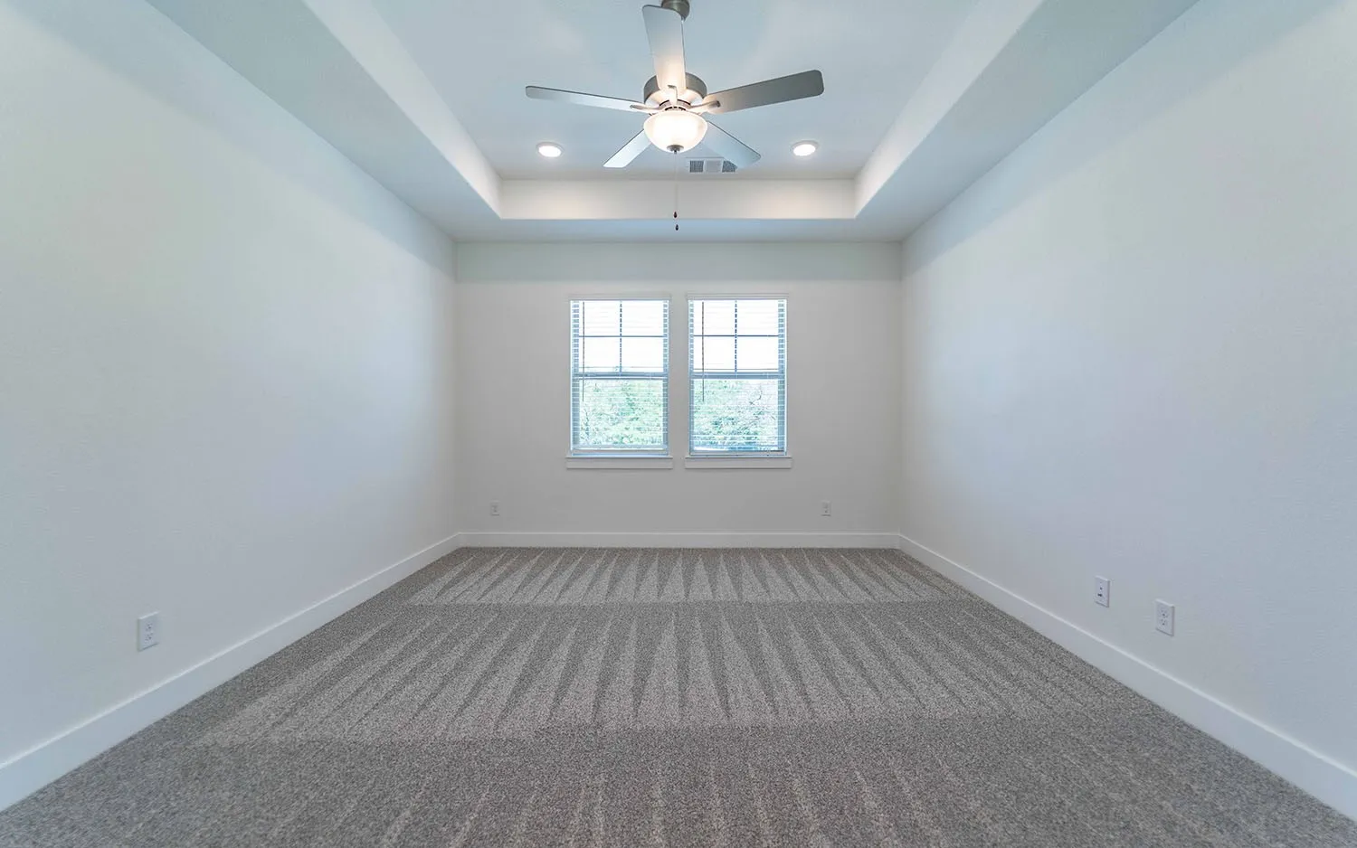 Spacious room featuring a tray ceiling with recessed lighting, a ceiling fan, plush patterned carpeting, and two windows with blinds