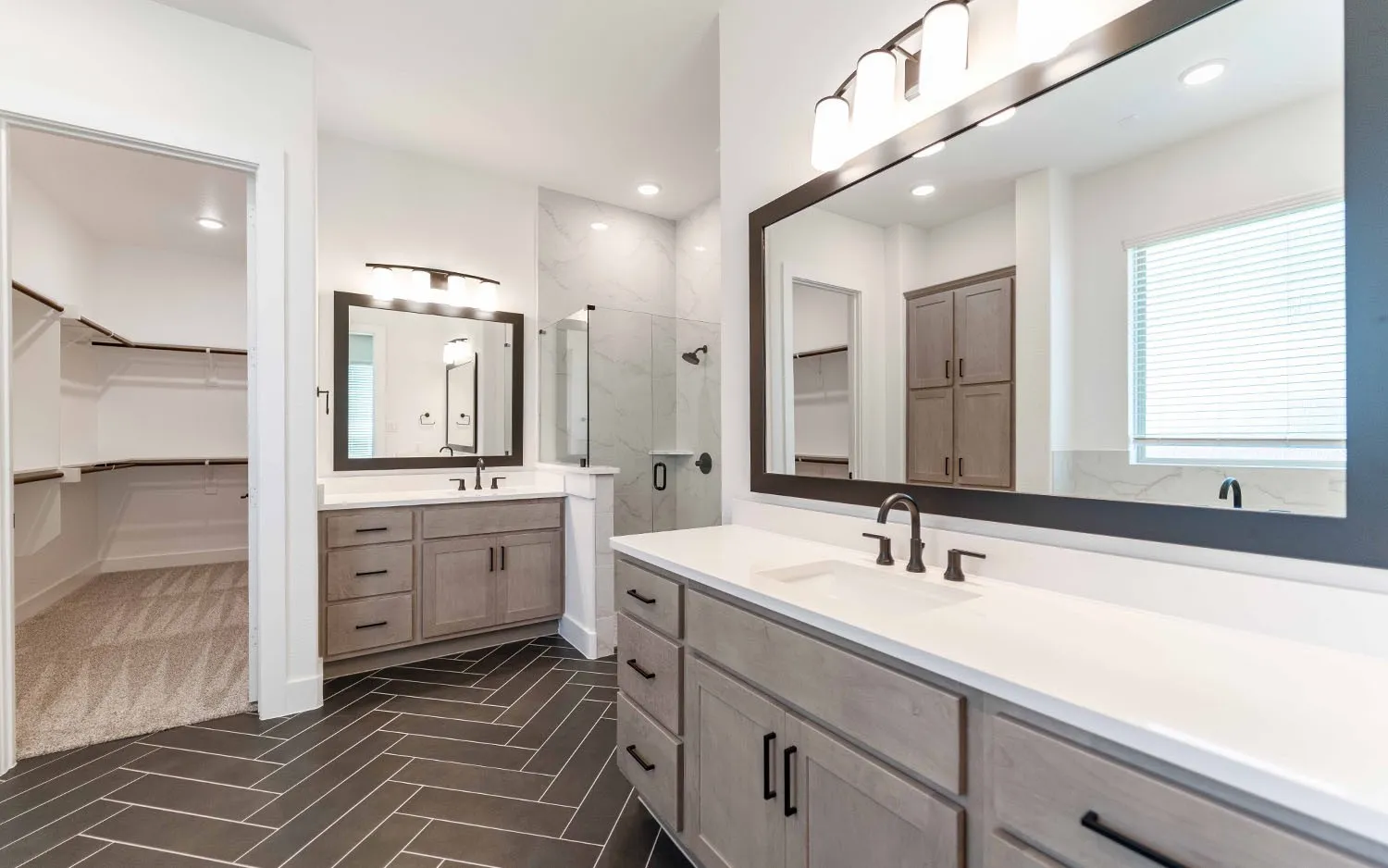 Spacious bathroom featuring dual vanities with white countertops and undermount sinks, herringbone-patterned floor tiling, a walk-in shower with a glass enclosure, and a large custom closet with built-in shelving